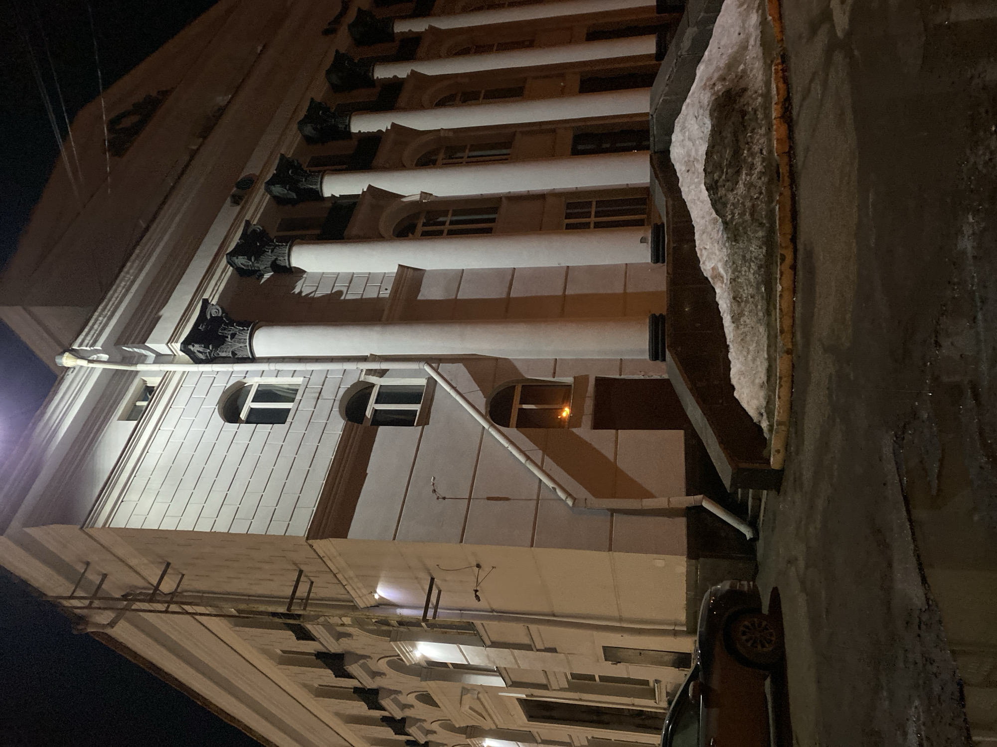 The image depicts the Perm Academic Opera and Ballet Theatre (Пермский академический театр оперы и балета им. П. И. Чайковского) at Petropavlovskaya Street, 25, Perm, Perm Krai, Russia, 614000, at night.
The scene captures the corner of a large, multi-story, classical-style building, predominantly cream or light beige, illuminated by artificial lighting under a dark sky. The architecture features numerous tall, white, fluted columns with dark, ornate Corinthian capitals extending upwards from a raised platform. The facade includes arched and rectangular windows, some with visible interior light. A prominent white downspout runs vertically along the left side of the building's corner, transitioning diagonally downwards. Metal ladder-like fixtures are secured to the upper section of the left wall. A dark-colored, possibly brown, entrance door is visible beneath an arched window displaying a warm, orange internal light source.
The ground in the foreground consists of dark, wet asphalt or concrete pavement, with visible puddles reflecting ambient light. A substantial mound of dirty, melting snow is accumulated on a raised curb or planter strip directly in front of the building's steps, indicating cold weather conditions. A dark-colored SUV or crossover vehicle is parked on the left side of the frame, with its front and driver's side partially visible. No individuals are present in the scene.