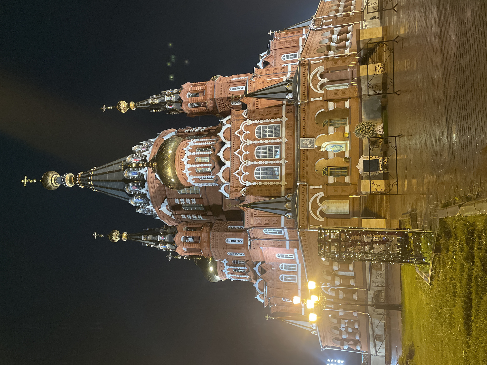 The image depicts the exterior of St. Michael's Cathedral in Izhevsk, Russia, at night. The cathedral is brightly illuminated, showcasing its red brick facade, white decorative trim, and multiple spires topped with golden domes and crosses. The surrounding area appears to be a paved surface reflecting some of the cathedral's light, indicating possible wet conditions. A section of lawn and a wrought-iron fence with string lights are visible. There are no people visible in the image.