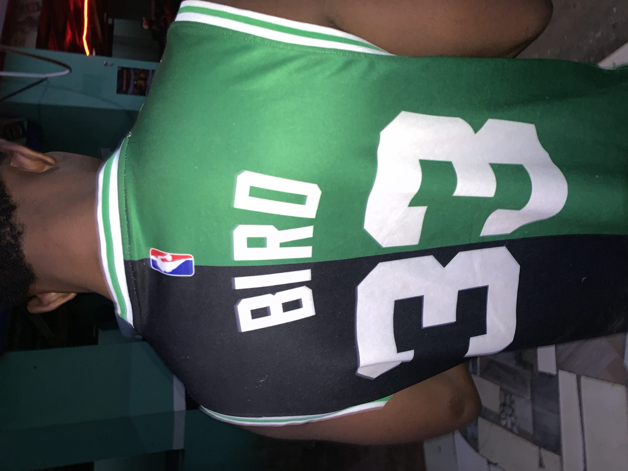A person wearing a Boston Celtics Larry Bird jersey with the number 33 is indoors. The jersey is green and black with white lettering and numbers. An NBA logo is on the upper left shoulder. The location is not New Stadium, Nigeria.