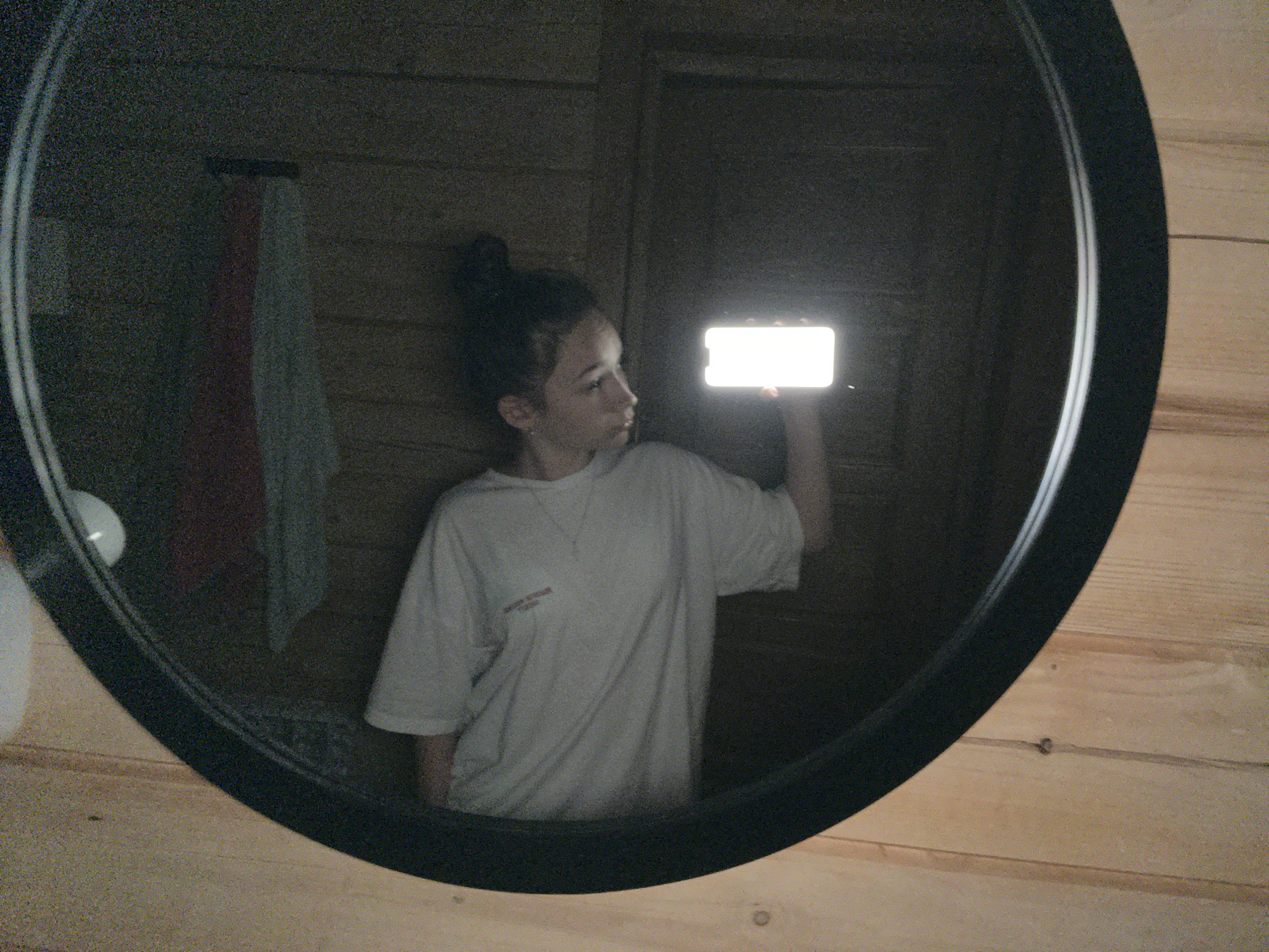 A person is taking a selfie in a circular mirror. The person is a young female with dark hair pulled up in a bun. She is wearing a light-colored t-shirt with a small, dark logo on the left chest. She is holding a smartphone horizontally in her right hand, and the screen is illuminated, acting as a light source for the photo. Her head is turned to the left, and she is looking at the phone.
The setting appears to be indoors, with wooden walls. The mirror is framed in black and is mounted on the wooden wall. In the reflection of the mirror, behind the person, there is a glimpse of what looks like a towel hanging on a rack, and a white object which could be a sink or a light fixture. The lighting in the scene is dim, with the primary light source coming from the illuminated phone screen. The overall impression is a casual, personal moment captured in a reflective surface. The time of day is indeterminate due to the indoor setting and artificial lighting.