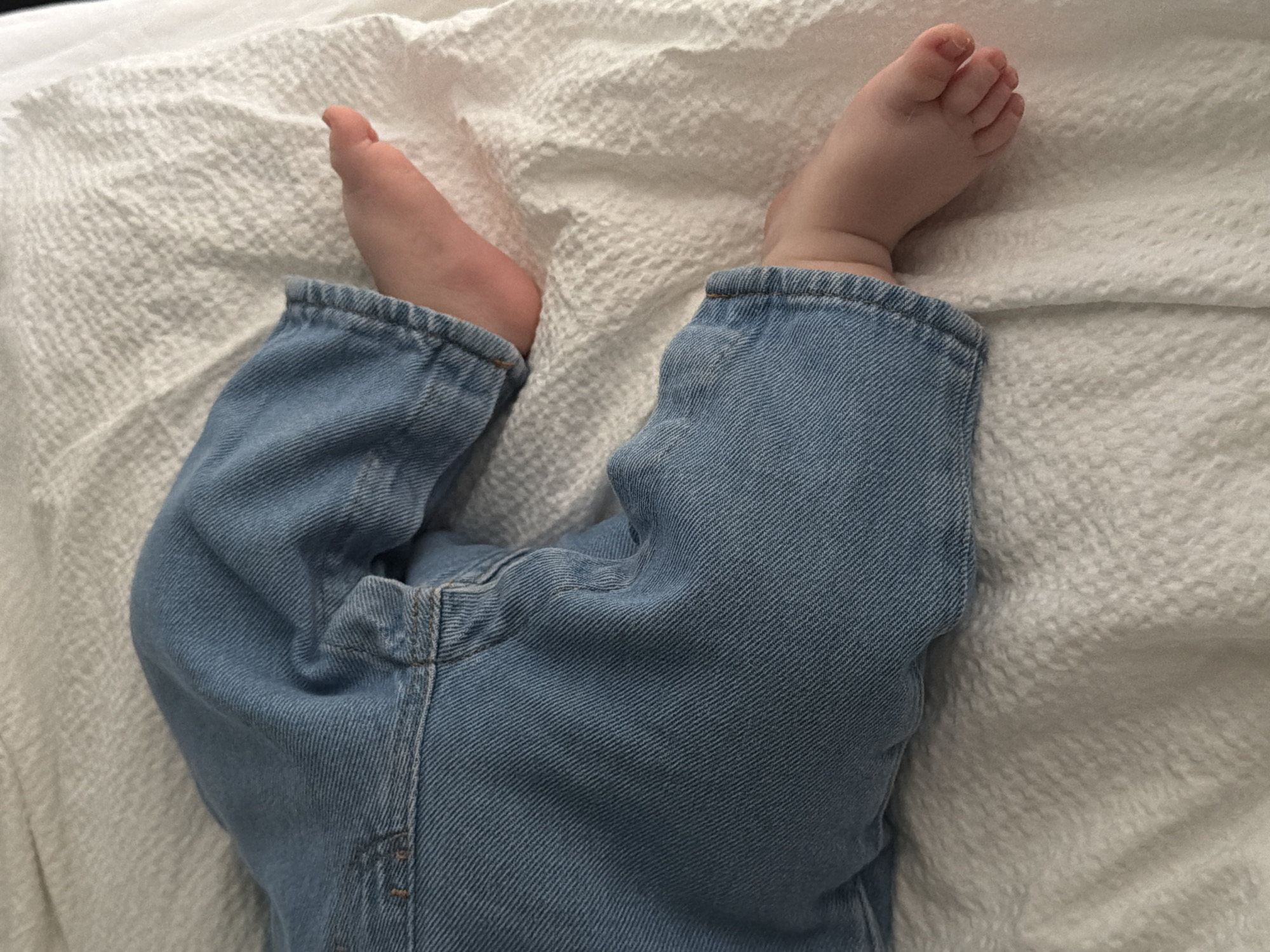 The image displays the lower half of a baby or toddler, focusing on their legs and feet, lying on a white, textured surface.
**Subjects:** The primary subject is a young child, evidenced by their small, bare feet. The child's body is mostly covered by clothing from the mid-calf downwards.
**Objects:** Key objects include a pair of light blue denim jeans worn by the child and a white, textured fabric (likely a blanket or sheet) that covers the surface beneath and around the child's legs. The jeans appear to be made of classic denim material, showing diagonal weave patterns and seams.
**Scene/Setting:** The scene is an indoor setting, likely a bed, sofa, or a cushioned surface, given the soft, textured fabric background. The child is positioned with their legs bent, and their feet are visible above the cuffs of the jeans. The general context suggests a calm, domestic environment in Khimki, Russia.
**Activity or event taking place:** The child appears to be resting or lying down in a relaxed state. Their feet are positioned casually, one pointing slightly upwards, indicating a lack of active movement or play.
**Notable details:** The child's feet are bare, pink, and small, with visible toes and arches. The jeans are a classic light blue wash, possibly a size larger than the child, as they gather around the ankles where the feet emerge. The white background fabric has a distinct raised, possibly waffle-like, texture, providing a soft contrast to the denim. There is no visible text in the image, nor any discernible cues regarding the time of day or specific weather conditions.