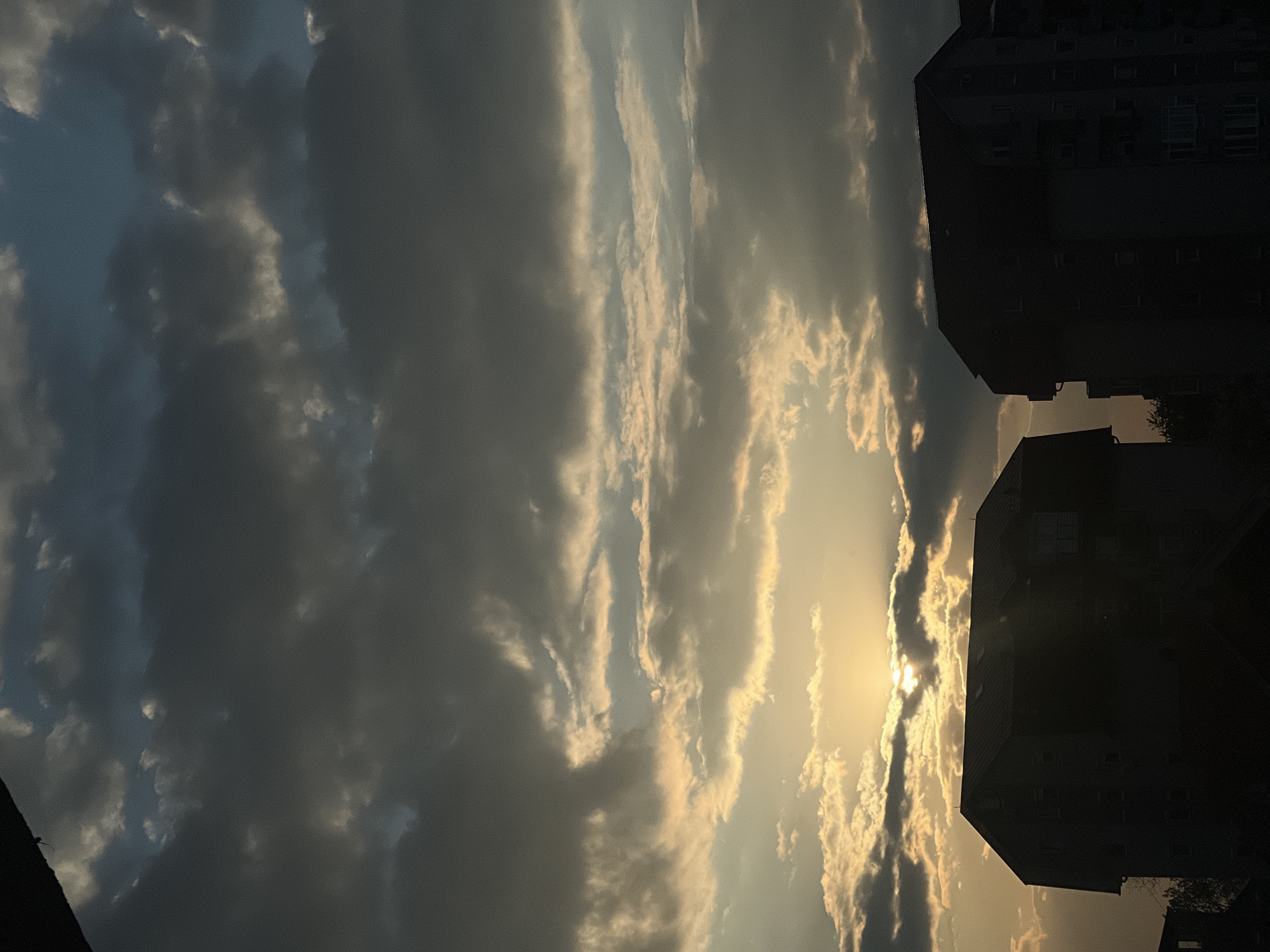 The image shows a section of the sky with the sun partially obscured by clouds, positioned over a building in Palilula Urban Municipality, Serbia. The clouds are a mix of light and dark gray, some appearing striated. Sunlight peeks through the cloud cover. The building on the right is dark, seemingly silhouetted against the bright sky. Details of the building's structure, such as windows, are faintly visible.