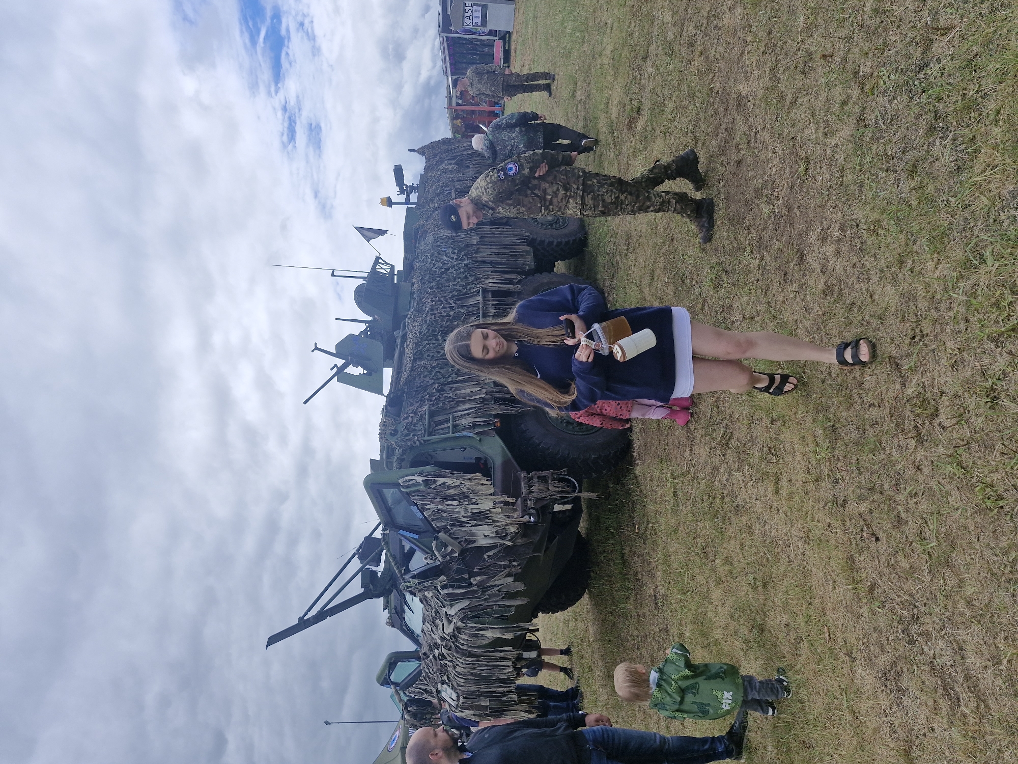 An outdoor scene captured under a cloudy sky depicts an event featuring military equipment on a dry, grassy field in Unknown, Unknown. A large, olive-green armored military vehicle, possibly a Patria Pasi or similar armored personnel carrier, dominates the midground. It is heavily draped with camouflage netting made of torn fabric strips, and its front two doors are open. The vehicle's roof features a weapon station, various antennas, and a yellow beacon light, with a small flag (white with a dark emblem) visible towards the rear.
In the foreground, a young woman with long brown hair, wearing a dark blue hooded sweatshirt, a white skirt, and black sandals, walks towards the left side of the frame. She is looking down at her phone held in her left hand and carries a white bucket containing a beverage container in her right hand. To her right, a man in a camouflage uniform with a dark beret and a blue sleeve patch walks in the opposite direction, smiling slightly.
To the left of the frame, a small child in a green hooded jacket with "PIX" written on the back stands facing the armored vehicle. A man in a dark jacket and jeans is partially visible further left. In the background, several other people, including individuals in military uniforms and civilians, are scattered across the field. A white trailer or container with "KASE" written on its side is visible in the far right background, adjacent to other temporary structures. The ground is covered with patchy, dry grass and exposed dirt, indicating foot traffic.