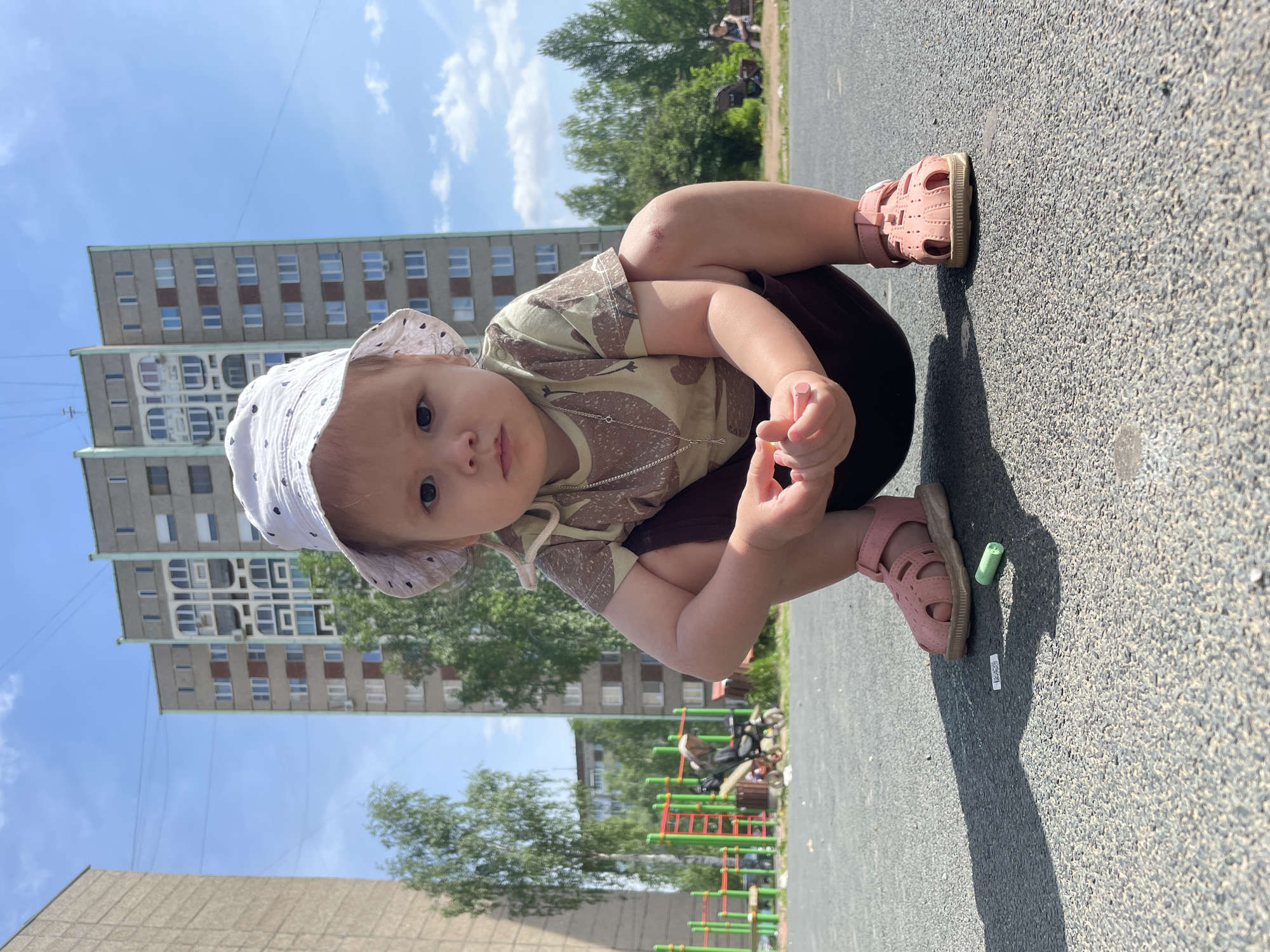 A young child in a white polka-dot hat, brown shirt, and pink sandals is squatting on a gray paved surface. The child is looking directly at the camera. The child's right foot is near a small green object and a white label on the ground. In the background, there is a tall, light-colored building with multiple windows, green trees, and a playground with green and red equipment. The location is Izhevsk, Russia.