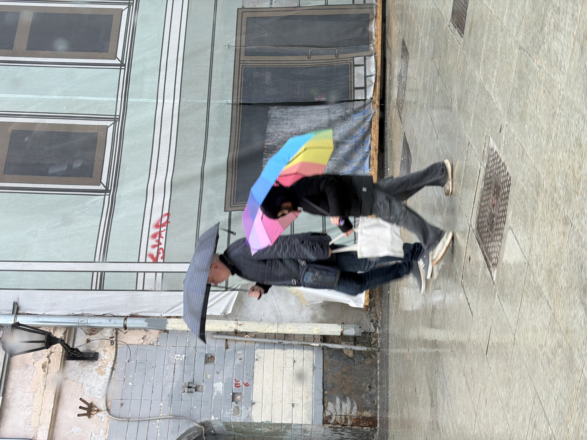 Two individuals walk under umbrellas on a wet, tiled sidewalk in Moscow, Russia. One person holds a colorful umbrella with pink, yellow, blue, and green panels. The second person has a grey umbrella. Both are dressed in dark clothing. They appear to be walking with purpose. The buildings lining the sidewalk show signs of wear, with faded paint, exposed brick, and painted murals. The murals depict windows and a door, creating an illusion on the building's surface. A vintage lamppost is affixed to one of the buildings. Red graffiti is visible near the murals.