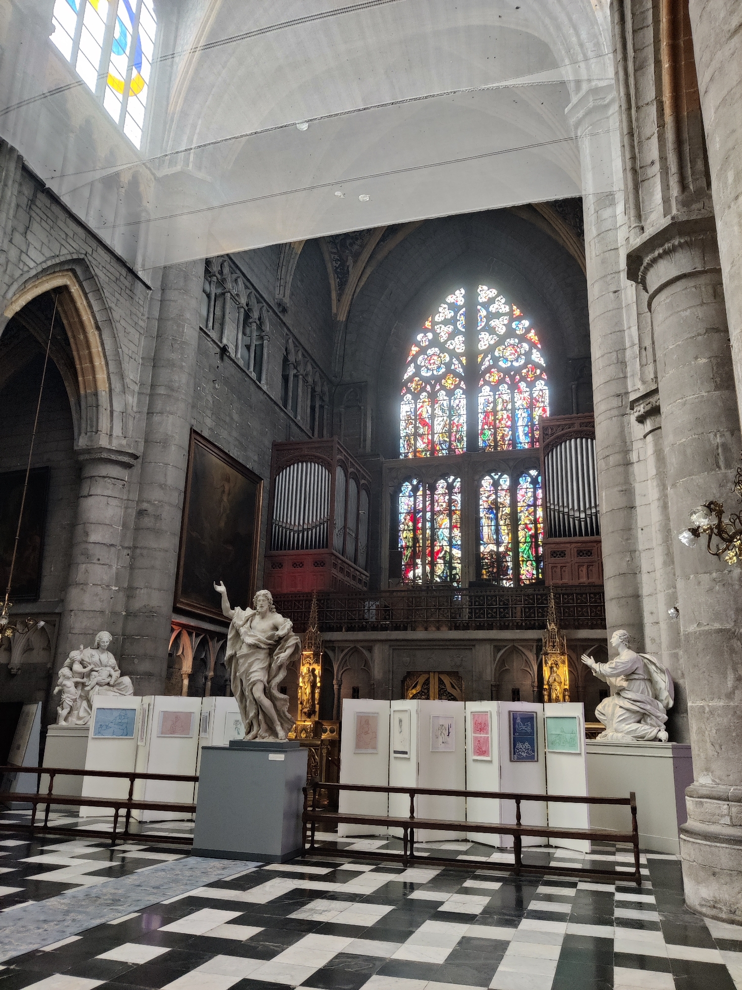 The image shows the interior of St. Paul's Cathedral in Liège, Belgium. The cathedral features a large stained-glass window behind the altar, with a pipe organ on either side. In the foreground, there are several white display panels exhibiting framed artwork. A statue of a man with his arm raised is positioned in front of the display panels. To the left of this statue, another white statue depicts a woman holding a child. The floor is tiled in a black and white checkered pattern. Large stone pillars and arched architecture are visible throughout the space, indicative of a historic church.