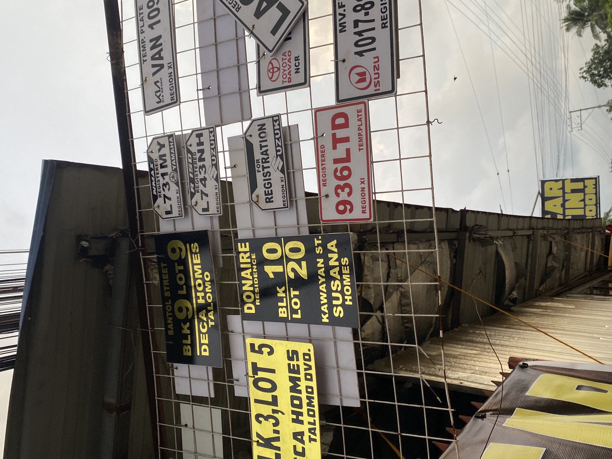 The image depicts an exterior scene in Davao City, Davao Region, Philippines, characterized by a display of various informational signs mounted on a white metal grid. The grid is suspended beneath a grey, weathered overhang, possibly part of a building or a large canopy structure.
On the left, prominent black and yellow signs advertise residential properties: one specifies "SANTOL STREET BLK 9 LOT 9 DECA HOMES TALOMO," and another, partially visible, reads "BLK. 3, LOT 5 DECA HOMES TALOMO DVO." Adjacent to these is a black and yellow sign for "DONAIRE RESIDENCE BLK 10 LOT 20 KAWAYAN ST. SUSANA HOMES."
Interspersed with these are numerous vehicle registration-related signs. These include white plates with black text, such as "REGISTERED L731MY NMAX YAMAHA REGION XI," "TEMP. PLATE KIA DAVAO VAN 109 REGION XI," "REGISTERED L743NH R150 SUZUKI REGION XI," and "FOR REGISTRATION SUZUKI REGION XI." A white sign with the red Toyota logo reads "TOYOTA DAVAO NCR." Another red plate with white text shows "REGISTERED 936LTD TEMP. PLATE REGION XI." A white plate with a red Isuzu logo has partially visible text including "MV.F 1017-8 ISUZU REGIS."
Below the grid on the left, a yellow and black banner is partially visible, displaying the text "CAR INT ROOM," suggesting a vehicle tinting service. The background shows an overcast sky with visible utility poles and numerous crisscrossing power lines. Sparse green foliage, including what appears to be a palm tree, is visible on the far right. A small, dark bird is captured mid-flight against the cloudy sky. The lower left portion of the image reveals a corrugated metal wall and a darker, sheltered area beneath the overhang. No individuals are visible in the frame.