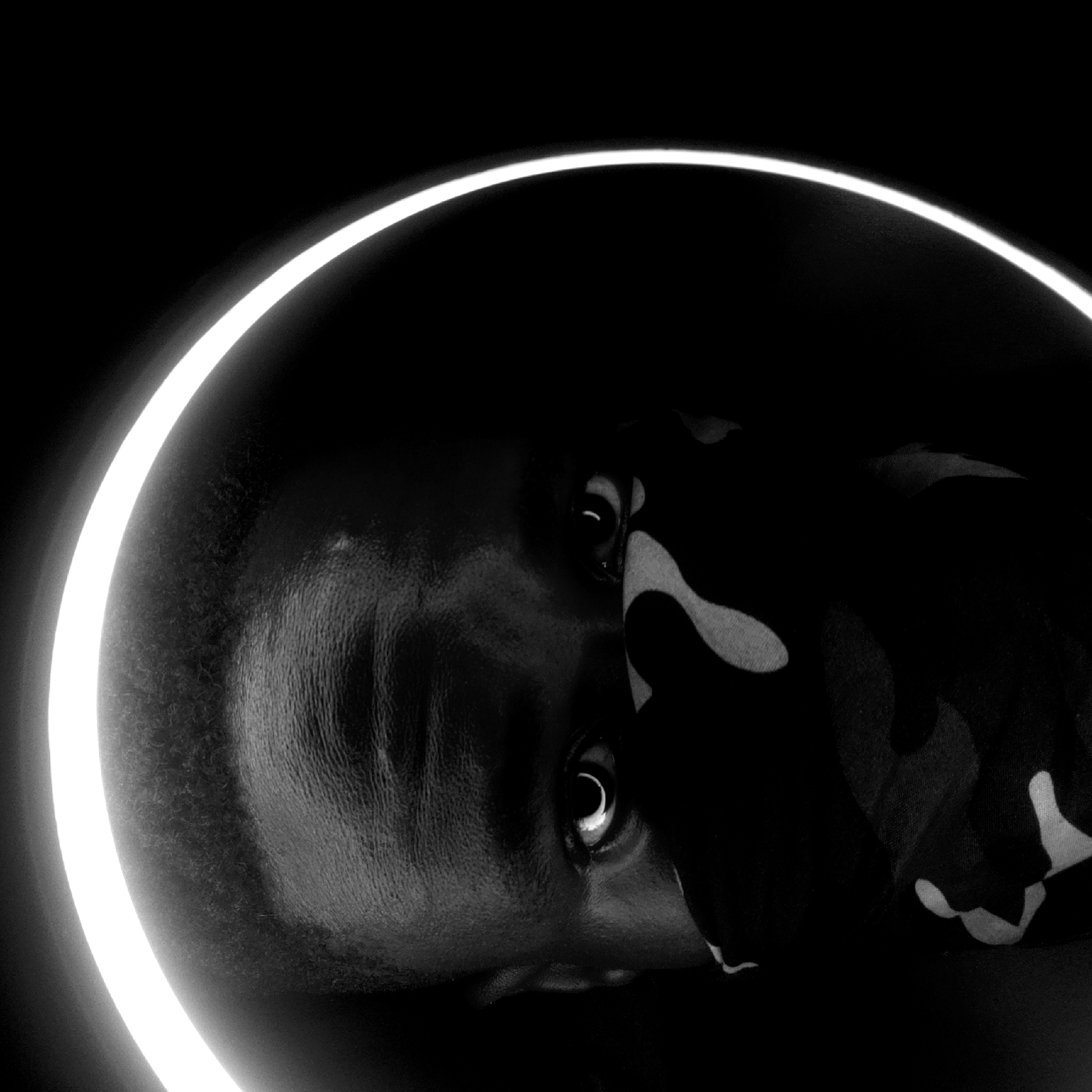 A black and white, close-up portrait of a person, likely male, with a partially obscured face. A light ring surrounds the person's head. The person's skin is dark and appears smooth. The person's eyes are visible. The person is wearing a patterned fabric covering part of their face. The image is dark, with the light ring providing the main illumination. There is no indication of being in Narayi, Nigeria.