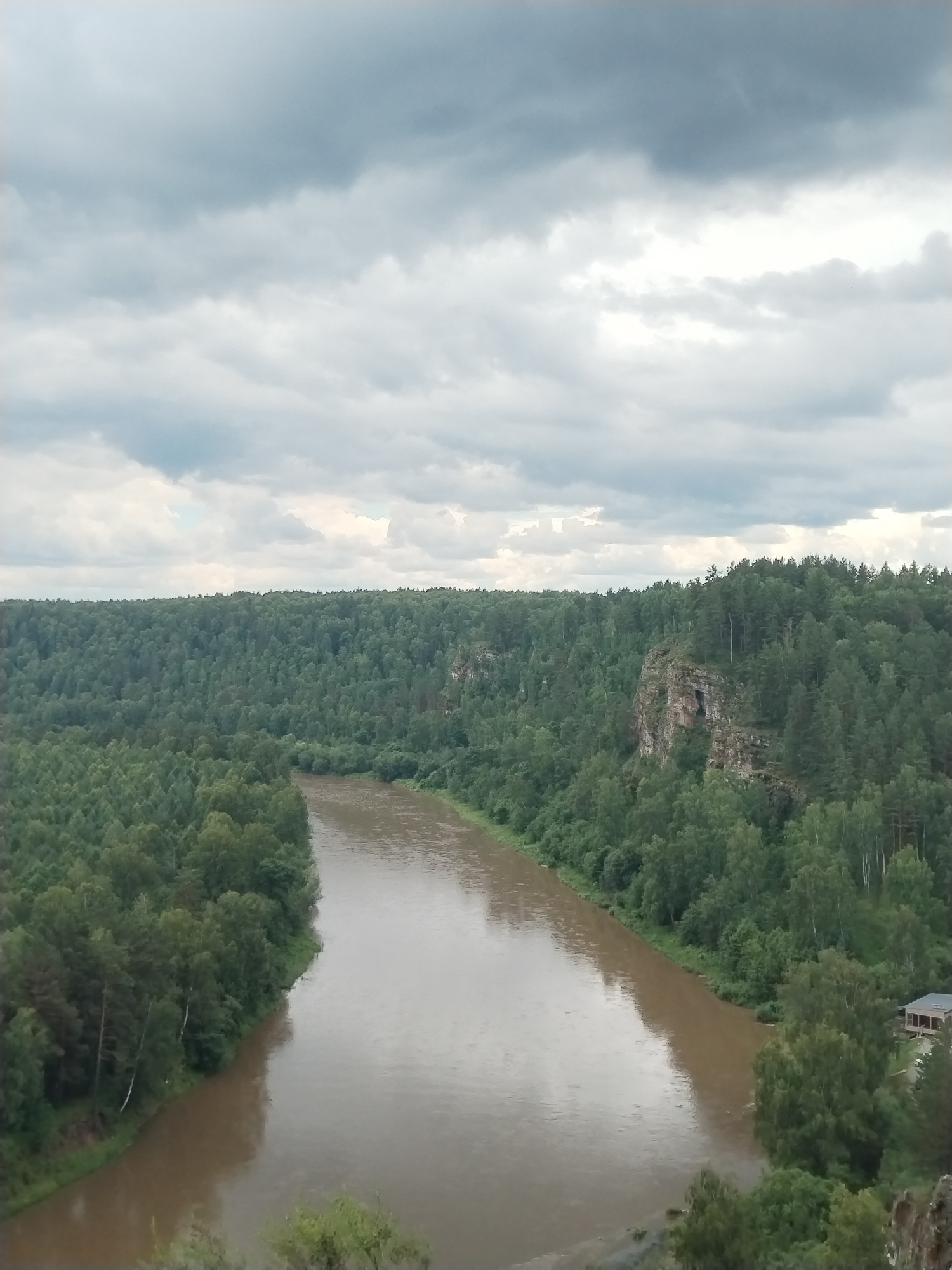 The image depicts a wide, muddy river flowing through a dense, green forest under a cloudy sky. The forest covers rolling hills, with a prominent rock formation featuring a cave visible on the right side of the frame. A small, wooden building with a dark roof is situated on the bank of the river on the right. The scene appears to be a natural landscape, likely in a rural or wilderness area.