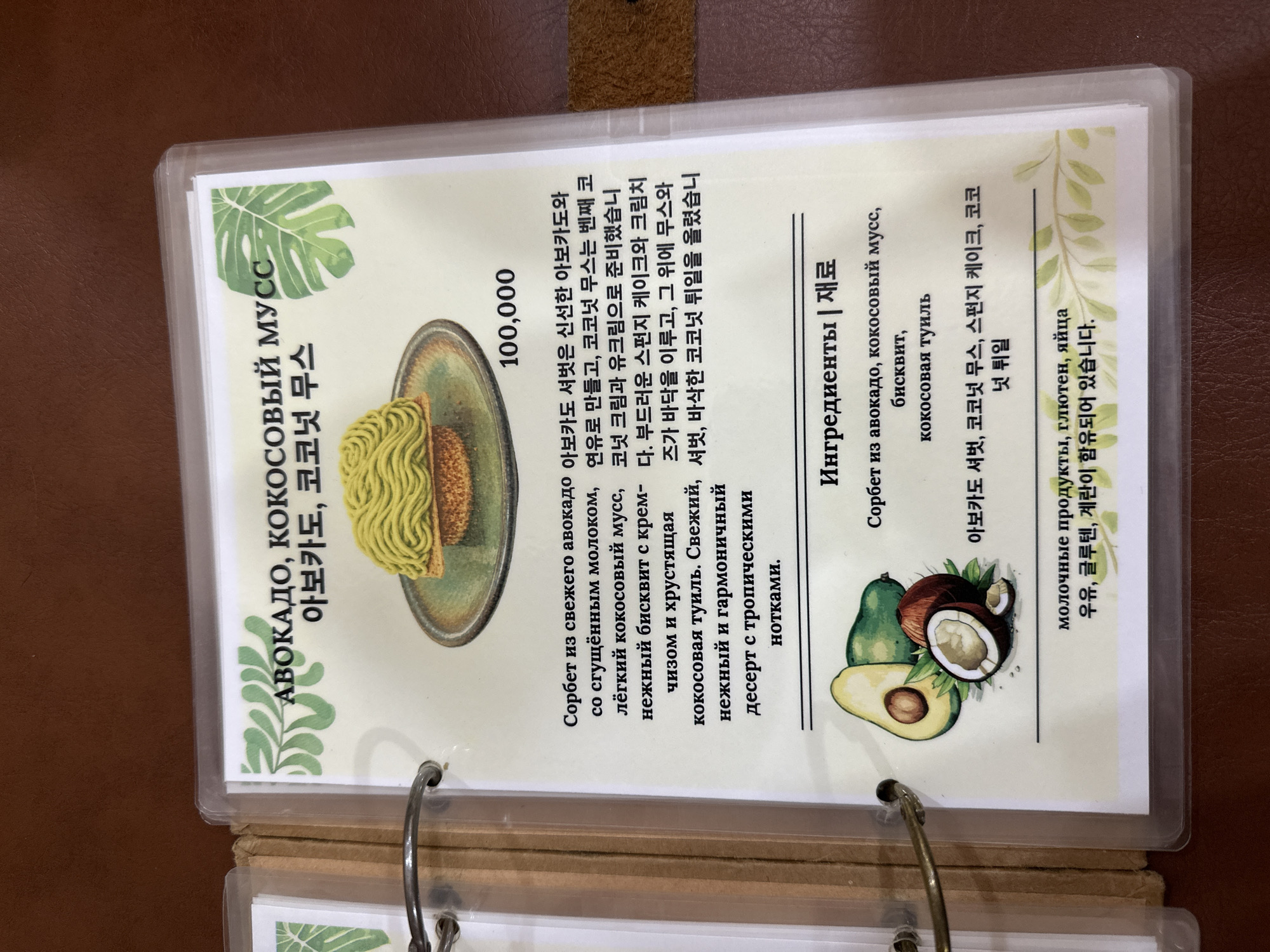 The image is a close-up of a menu page, likely from a cafe or restaurant, with text in both Russian and Korean. The menu item features an avocado and coconut mousse dessert, described as a sorbet made with fresh avocado and condensed milk, light coconut mousse, and a soft biscuit with cream cheese and crunchy coconut tuiles. The dessert is topped with a green, swirled mousse. The price listed is 100,000. Beneath the dessert description are ingredients, including avocado, coconut mousse, biscuit, and coconut tuiles. A separate section indicates that the product may contain dairy, gluten, and eggs. The overall presentation suggests a visually appealing and possibly gourmet dessert. The background of the menu page has a light, textured pattern with green leafy illustrations. The surrounding environment appears to be a table with a brown, textured surface. The image does not provide any information about the city of Nha Trang, Vietnam.