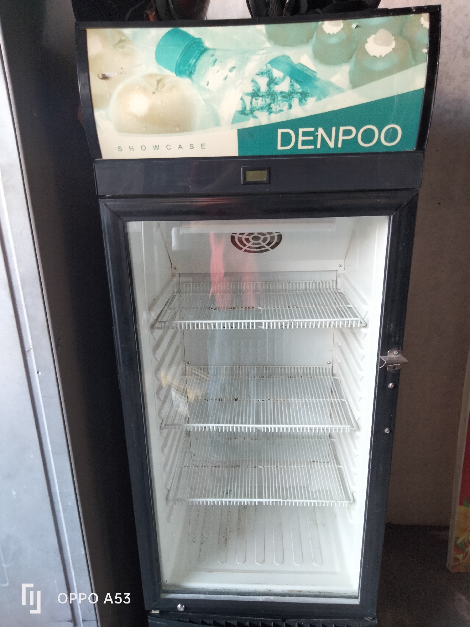 A commercial display refrigerator with the brand name "DENPOO" is pictured. The refrigerator is empty, featuring three wire shelves and a fan vent at the top. A bottle opener is attached to the right side of the refrigerator door. The image also includes a partial view of another appliance to the left, possibly a refrigerator or freezer, and a wall in the background. The text "OPPO A53" is watermarked in the lower left corner, indicating the device used to capture the image. The scene appears to be indoors, likely in a commercial or retail setting.