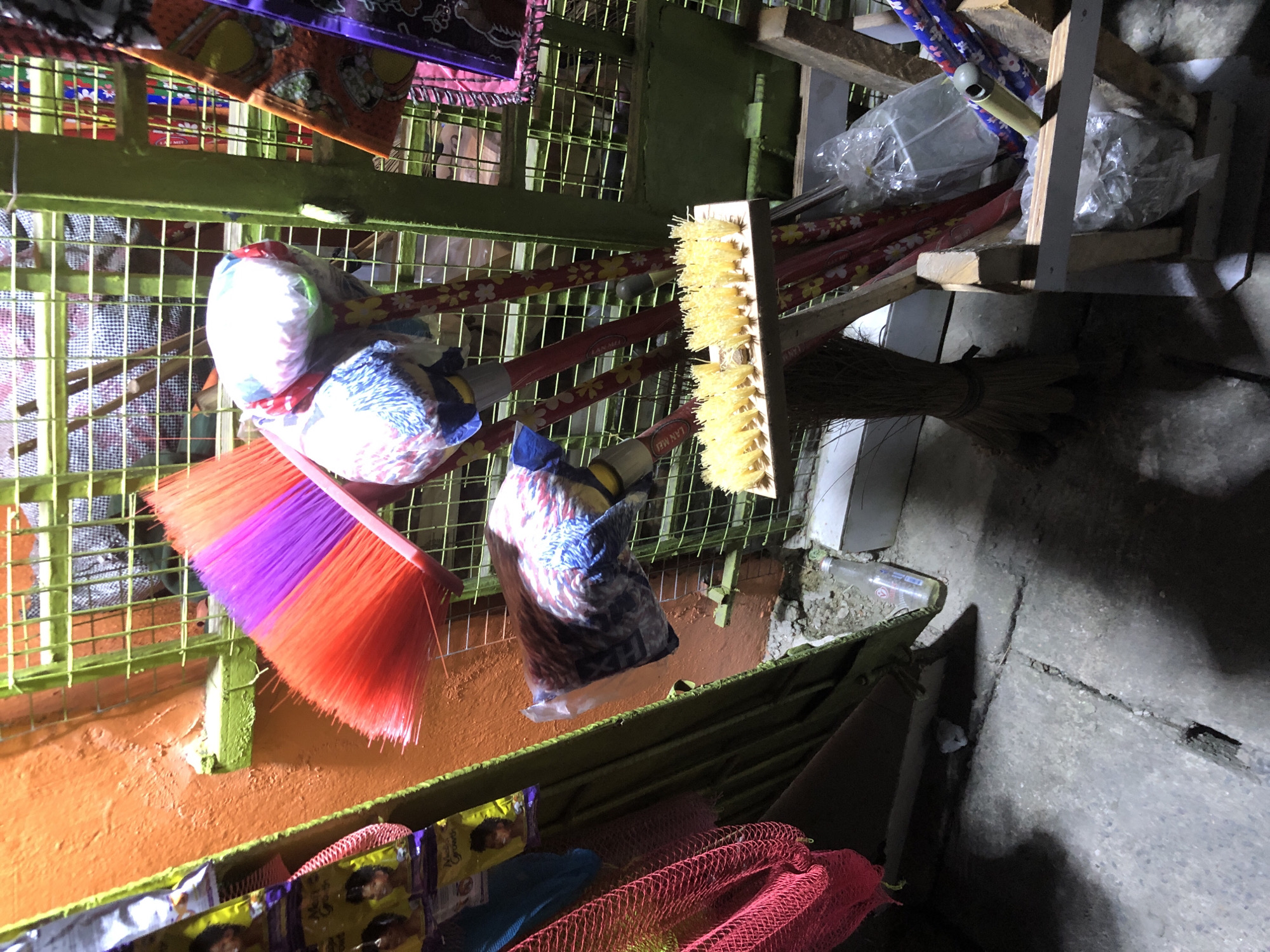 This image captures a storefront display in Ado, Nigeria, featuring various cleaning supplies. Dominating the foreground are several brooms, one with bright red and purple bristles, and another with a yellow brush head, propped up on wooden stands. Other brooms with decorative red handles are visible, some wrapped in plastic. Behind the brooms, a green metal grate encloses the display, revealing more items, possibly additional cleaning tools or household goods, within the store. Adjacent to the display is an orange-painted wall, and to the left, a rack holds packages of what appear to be hair products, some featuring images of women. The ground is concrete with visible cracks and a small grate. A clear plastic bottle is also present on the ground. The lighting suggests it is nighttime or a dimly lit area, casting shadows.