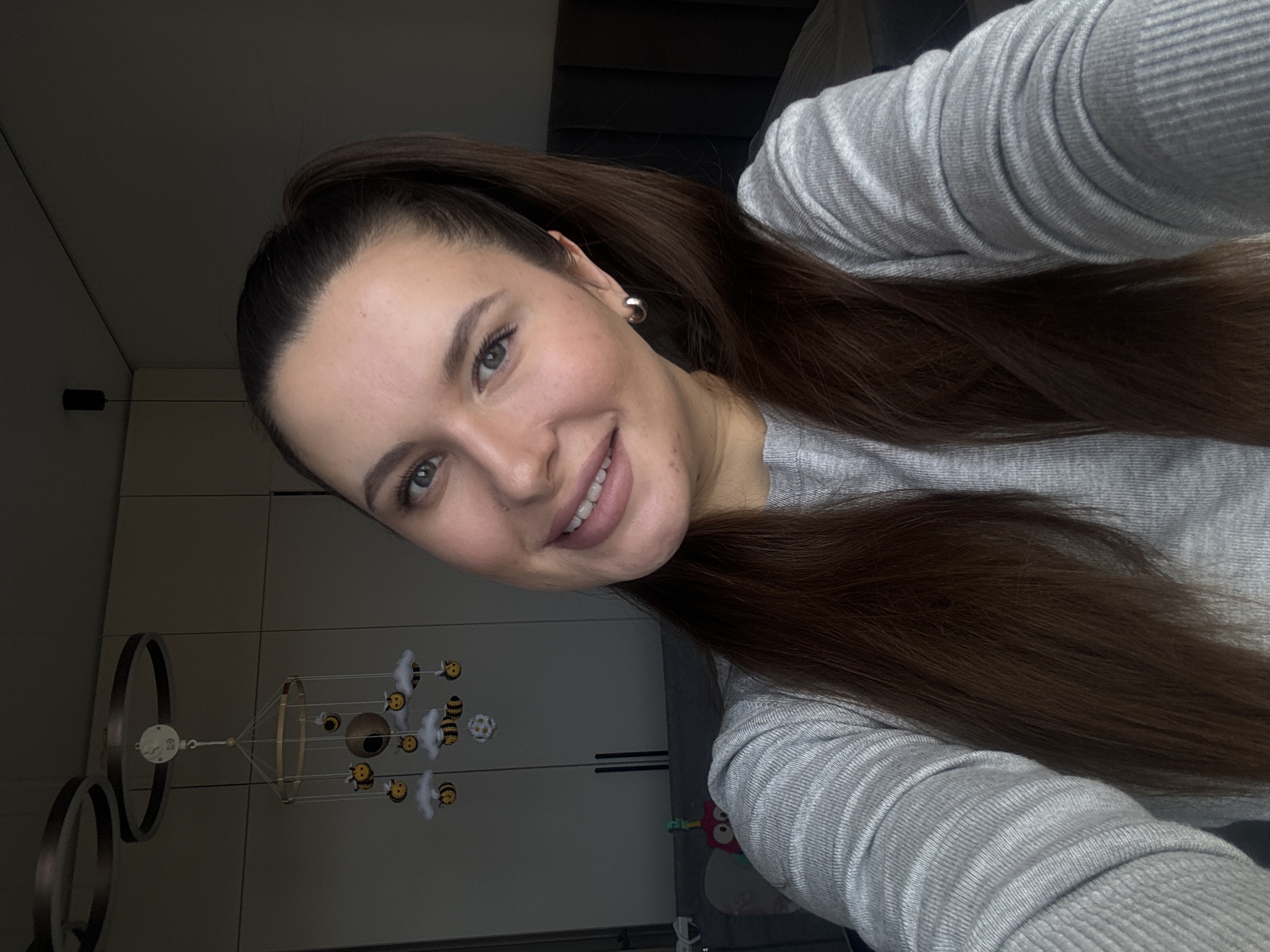 The image is a selfie of a woman, oriented vertically (rotated 90 degrees counter-clockwise), taken indoors in what appears to be a home in Akulovo (Gp Kubinka), Russia.
The primary subject is a woman with long, dark brown hair pulled back in a high ponytail. She is looking directly at the camera with a friendly smile, revealing light pink lips and white teeth. Her eyes are light-colored, possibly green or hazel. She is wearing a light grey, ribbed long-sleeve top and a silver hoop earring in her visible left ear. Her complexion appears clear, with slight blush on her cheeks.
The background suggests a domestic setting. To her right (image left), a modern light fixture with two concentric dark rings is partially visible. Centered in the background is a child's mobile hanging from the ceiling, featuring small yellow and black bees, white clouds, and a central brown sphere. Behind the mobile, large, plain white or light-colored wardrobe doors with dark horizontal handles can be seen. The overall lighting in the room is soft, suggesting natural light from a window not directly in view or ambient artificial lighting. No discernible text is visible in the image. The woman's expression is one of contentment or happiness.