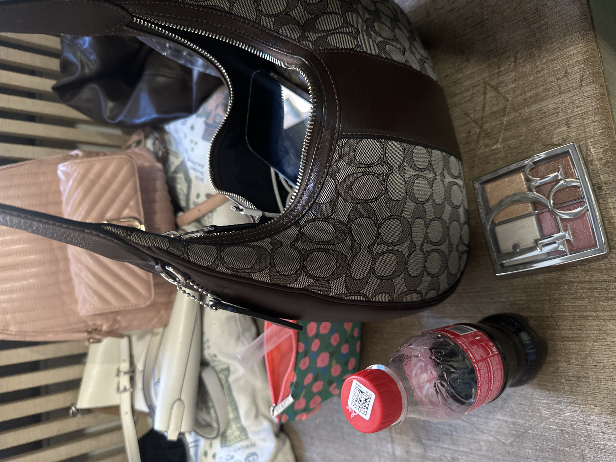 The image depicts a collection of personal items on a wooden surface. A partially open brown Coach handbag is prominently positioned. Inside the handbag, other items are visible, including a light pink quilted purse and what appears to be a smartphone. To the right of the handbag is a Dior makeup palette. Adjacent to that is a partially consumed Coca-Cola bottle with a red cap. The scene provides insufficient data to identify Yerevan, Armenia as the location. No information to suggest the subject is in the city of Yerevan, Armenia. The wooden surface the objects rest on appears to be slatted. Other bags and wallets are also in the image.
