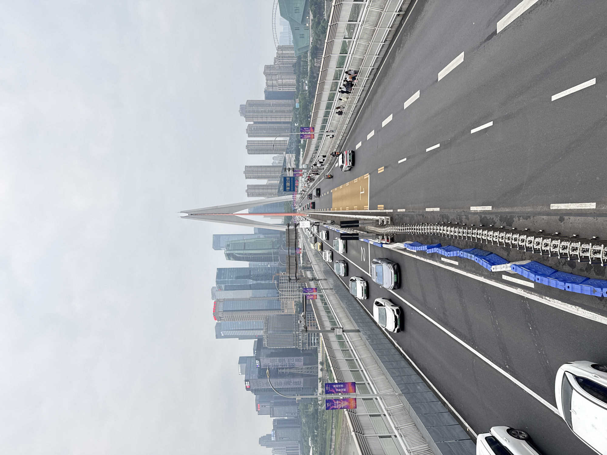 The image captures a perspective from a busy bridge in Yuzhong District, China, overlooking a cityscape dominated by modern skyscrapers. The bridge itself features multiple lanes of traffic with cars of various colors and models moving in both directions. Pedestrians can be seen walking on a designated pathway along the side of the bridge, separated from the vehicular traffic by a railing. A prominent cable-stayed bridge tower rises in the background, with its distinctive white structure piercing the overcast sky. Banners with Chinese characters and colorful graphics are visible along the bridge's edge, likely advertising local events or businesses. The urban environment is dense, with numerous buildings of varying heights and architectural styles forming a panorama across the horizon. The overall atmosphere is one of a bustling metropolitan area on a cloudy day.