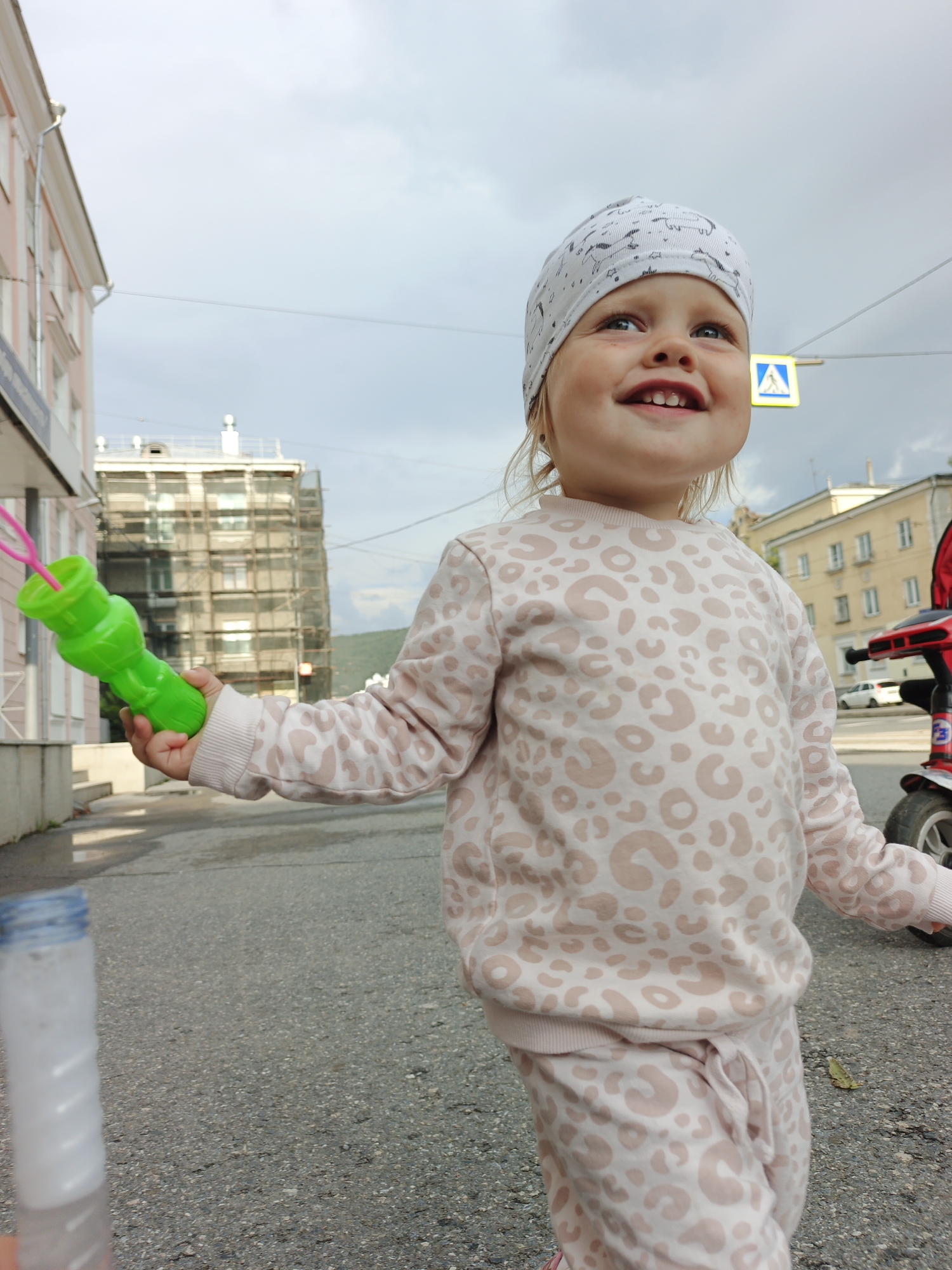 A young child is standing on a paved street, holding a green bubble wand with pink loops. The child is wearing a white bandana with a black pattern and a light pink sweatshirt and pants with a leopard-like print. The child is looking up and to the right, with a smile. In the background, there are buildings, a street sign, and a portion of a red tricycle. The sky is overcast. The image appears to have been taken in Zlatoust, Russia, given the architectural style of the buildings and the presence of a Russian street sign.