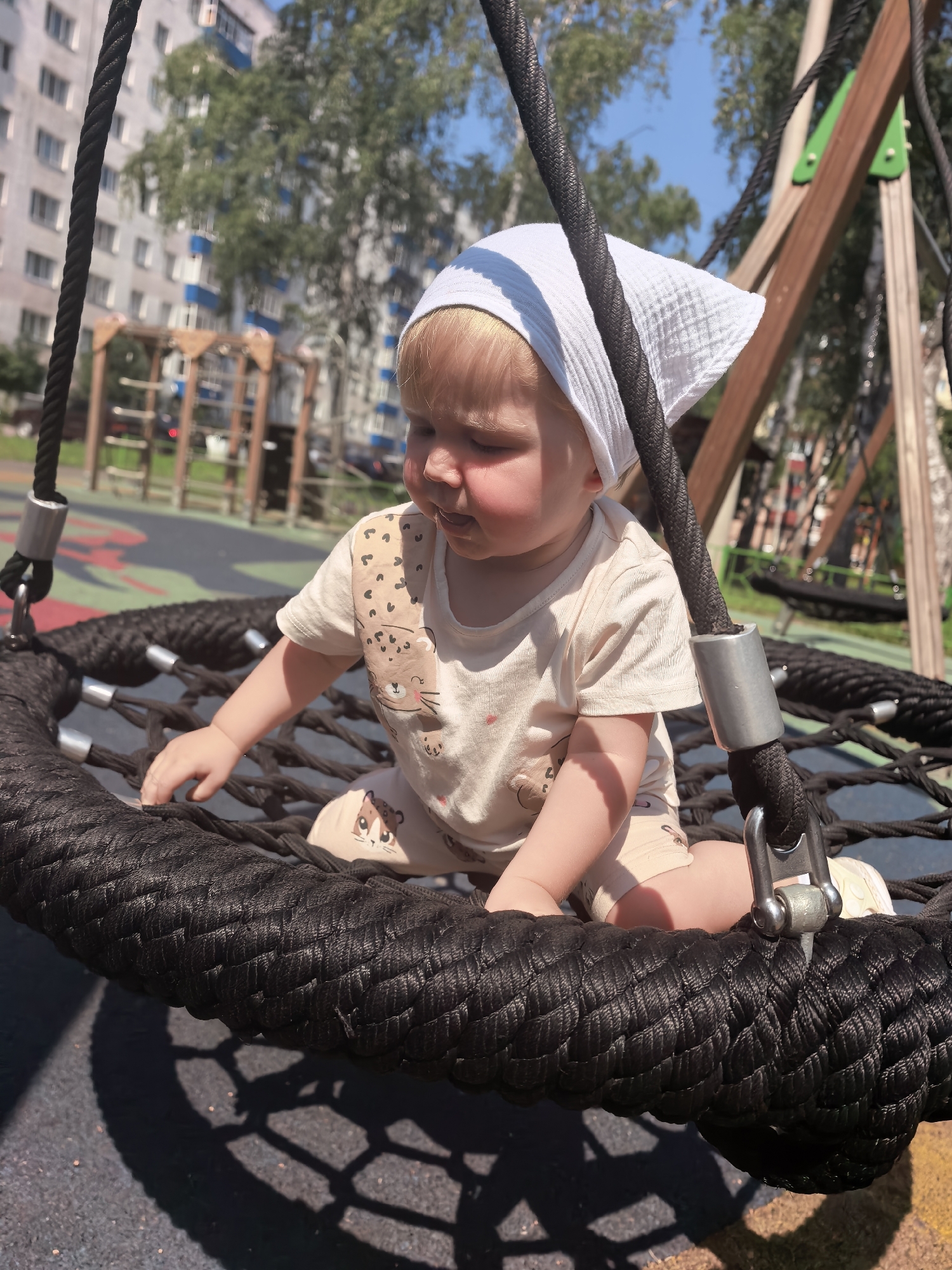 A young Caucasian child with blonde hair, wearing a white headscarf and a light-colored t-shirt with a leopard print, sits on a black rope swing at a playground. The child's face is turned downwards, and their right hand is reaching towards the rope of the swing. The swing is suspended from a wooden structure, and the ground beneath appears to be a soft, multi-colored surface typical of a children's play area. In the background, there are trees and a tall, light-colored apartment building with blue accents. The sky is clear and blue. This scene is located in the city of Sterlitamak, Russia.