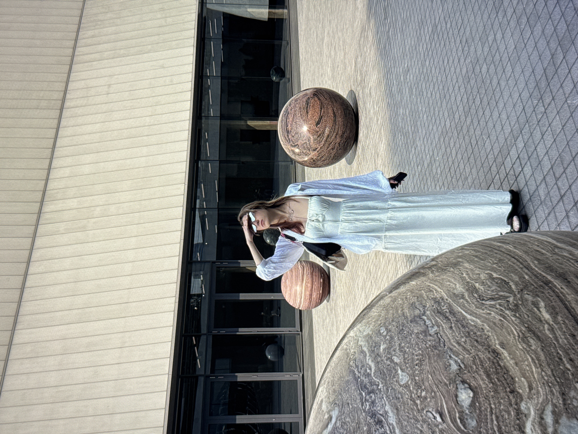 A young woman in a light blue dress and a white open cardigan stands on a paved plaza, shielding her eyes from the sun with her hand. She wears sunglasses and a black purse is slung over her shoulder. In the background, a modern building with a textured facade and large glass doors is visible. Several large, spherical sculptures are scattered across the plaza; one in front of the building appears to be made of marble with swirling patterns, and another to the right is a reddish-brown sphere with a similar marbled texture. The scene is set during the daytime under a clear sky. This location is the National Research University Higher School of Economics (HSE) campus on Myasnitskaya Street in Moscow, Russia.