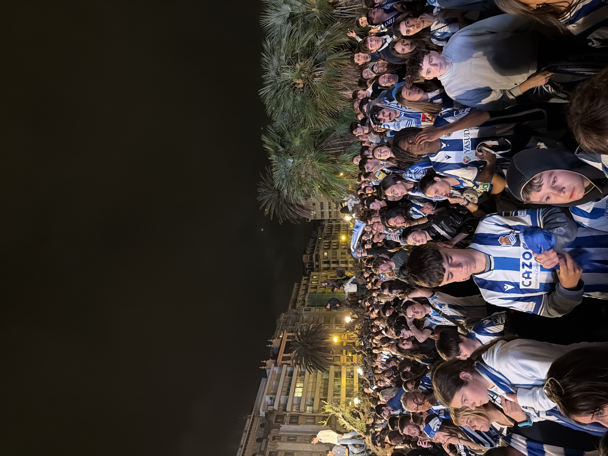 The image captures a large crowd of people gathered on a street in San Sebastián, Spain, likely during an evening event. The crowd is densely packed, with individuals wearing blue and white striped clothing, suggesting they may be supporters of a local sports team. Palm trees are visible, interspersed among the crowd and along the edge of the street, adding to the scenery. Buildings, illuminated with lights, line the street, creating a nighttime urban backdrop. The sky above appears dark, indicating it is night. The crowd appears to be focused on something, though the exact nature of the event is not clear from the image.