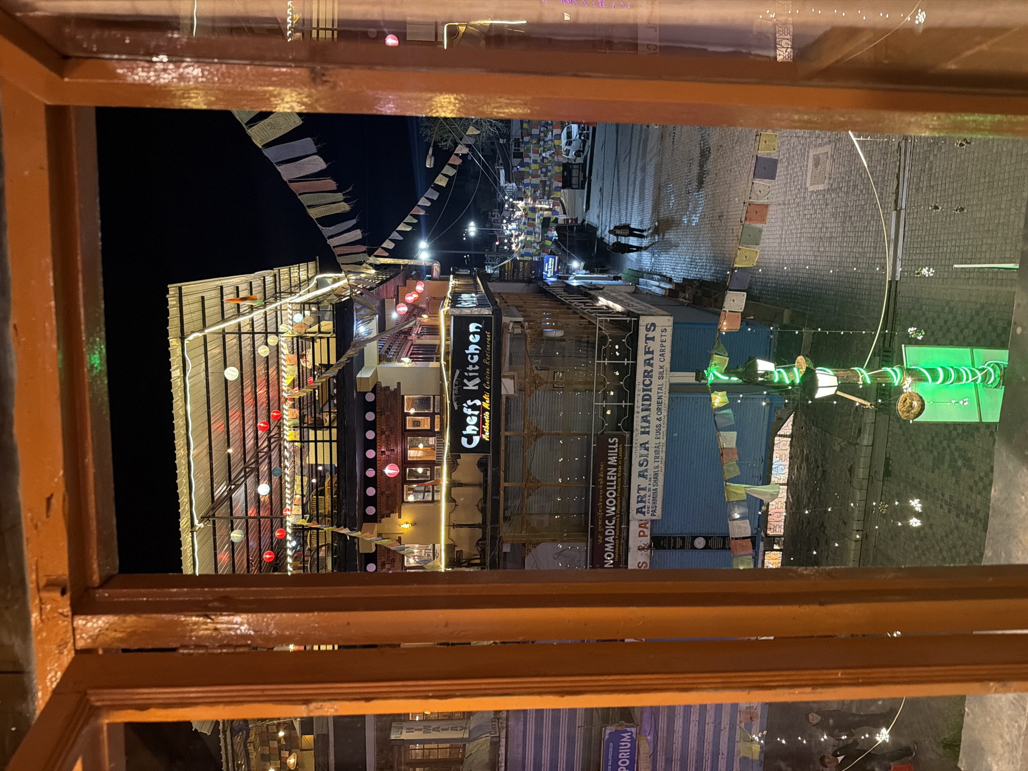 This image provides a view of a street scene at night in Leh, India, seen through a wooden window frame. The street is paved with stone and appears wet, reflecting the lights from the surrounding buildings and overhead decorations.
In the foreground, the warm orange-toned wooden frame of a window partially obscures the view. The frame has some texture and appears worn in places.
Dominating the mid-ground is a multi-story building with multiple levels. The uppermost level features a covered outdoor dining area with a metal roof, illuminated by strings of white lights and numerous decorative paper lanterns in red, white, and green. Below this, other levels of the building have windows and signage. One prominent sign reads "Chef's Kitchen" in white lettering on a black background, with smaller text below indicating "Authentic Multi Cuisine Restaurant". Other signs visible include "NOMADIC, WOOLLEN MILLS" and "S & PA ART ASIA HANDICRAFTS". Prayer flags, in various colors, are strung across the street and between buildings, a common sight in the region.
The street itself is lined with shops and businesses, many of which are lit up. There are several lampposts, one of which is notably decorated with green LED lights. Small strings of white lights are also draped along some buildings and the street.
In the background, the street extends into the distance, with more buildings and lights visible. Several vehicles, including a white van, are parked or moving on the street. Two individuals are walking on the wet pavement in the mid-ground, appearing to be heading away from the viewer. Another person is seen walking on the sidewalk to the left, near a sign for "EMPORIUM".
The overall atmosphere is lively and festive, with the night lights creating a warm glow on the scene. The wet pavement suggests recent rain or that the street is being cleaned. The presence of prayer flags and the architectural style of the buildings are indicative of Leh, a city in the Ladakh region of India known for its Tibetan Buddhist culture.