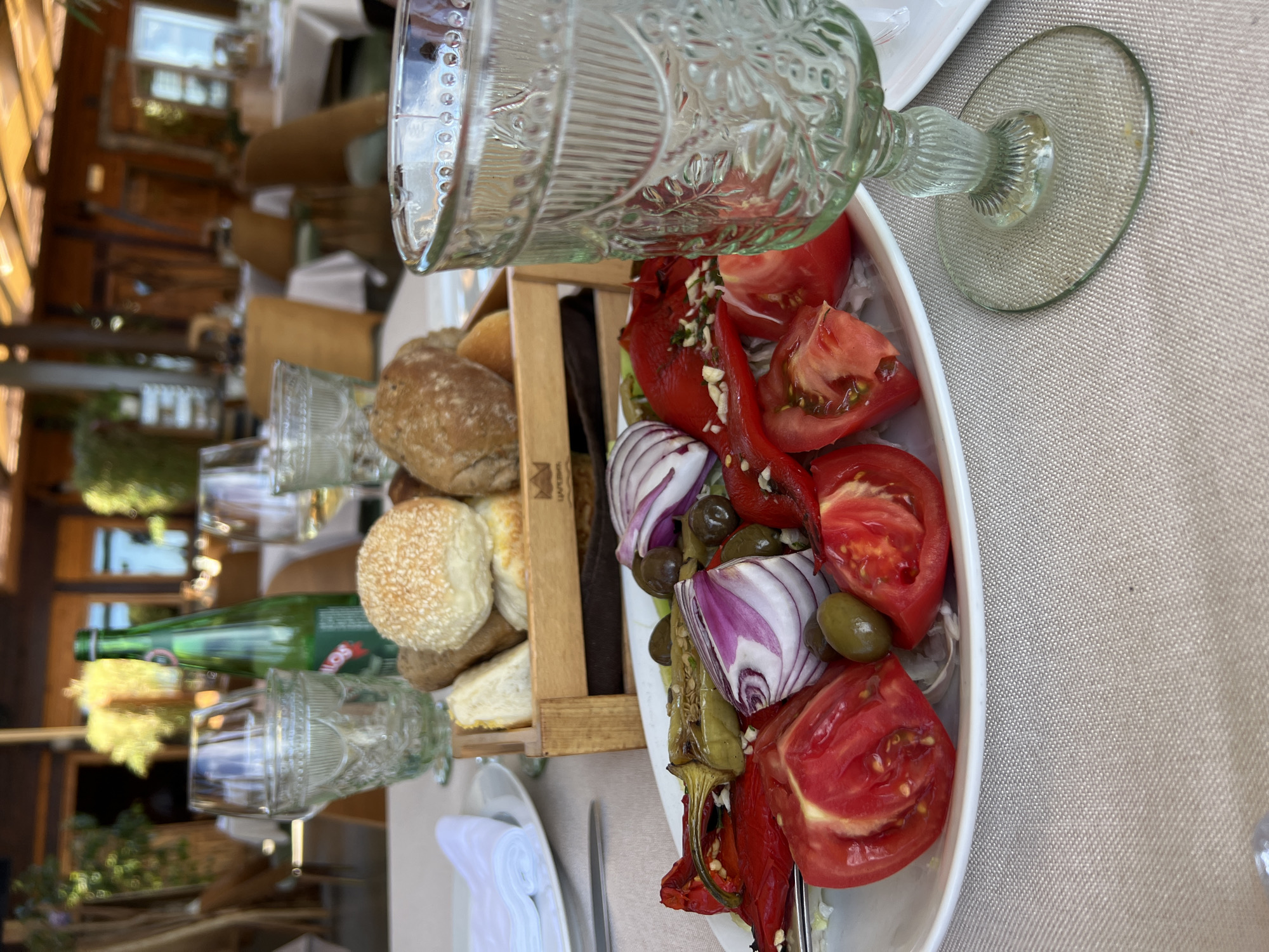 The image shows a close-up of a table setting at an outdoor restaurant, likely in the city of Kovačko Polje, Montenegro. In the foreground, a white plate holds a colorful salad composed of sliced tomatoes, red onion wedges, olives, and grilled green peppers. Behind the salad, a wooden crate contains a selection of bread rolls, including one topped with sesame seeds. To the left, a white napkin is neatly folded on a white plate, and a silver fork rests beside it.
Dominating the right side of the frame is a decorative, clear glass with a textured pattern, positioned on a table covered with a beige tablecloth. Further back, several other glasses and a green bottle of water are visible. The background reveals the restaurant's ambiance, with blurred images of wooden chairs, tables with white tablecloths, and overhead wooden beams, suggesting an open-air dining area. The overall scene depicts a meal ready to be served in a pleasant, possibly Mediterranean, setting.