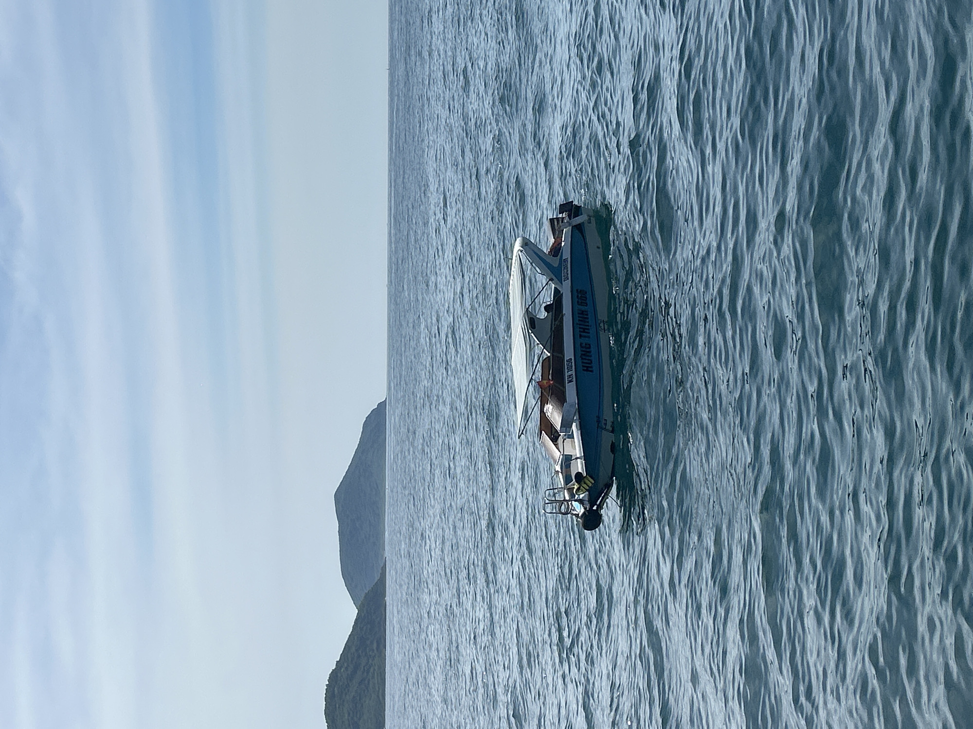 A small motorboat with a white canopy is floating on the calm, blue sea. The boat has blue and white trim, and the text "HUNG THINH 868" is visible on its side. Two people are seated inside the boat. In the background, lush green islands rise from the water, with a hazy sky above. The scene appears to be the coast of Nha Trang, Vietnam.