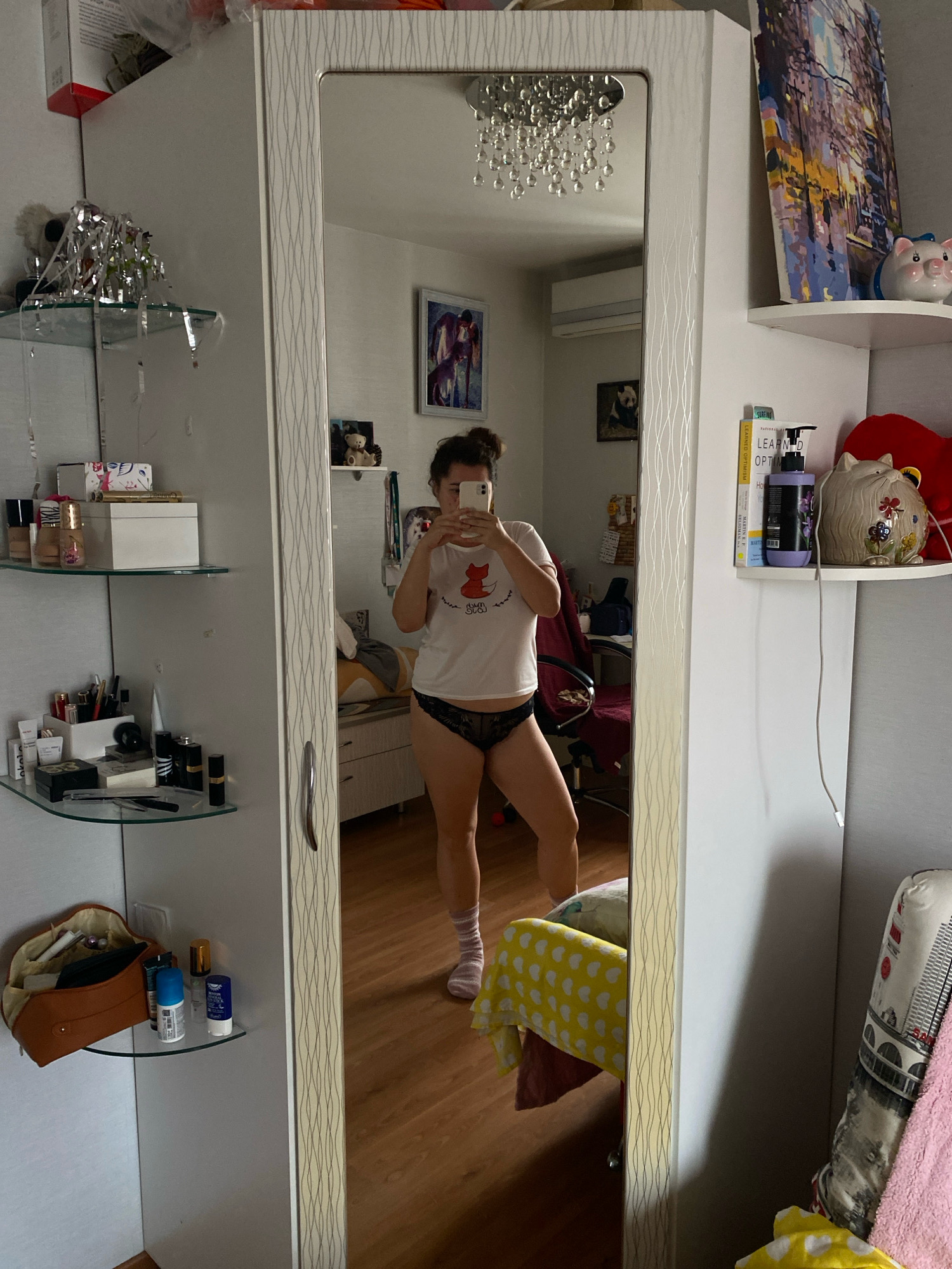 A person stands in front of a mirror, taking a selfie. They are wearing a white t-shirt with a red fox graphic and black lace underwear. The room appears to be a bedroom, with a bed visible in the reflection of the mirror. There are shelves on the wall filled with various items, including toiletries, books, and decorative objects. The overall scene suggests a casual, personal moment captured in a domestic setting. The location is not specified in the image or crops.