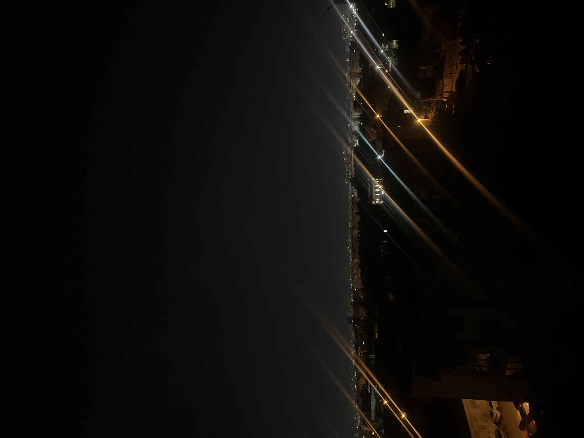 The image depicts a nighttime view of what appears to be a coastal city from an elevated vantage point. The city is lit by streetlights and building lights, which create streaks of illumination. A large body of water, possibly the sea, is visible on the left side of the frame. Buildings, streets, and other urban features are discernible along the coastline. The scene is predominantly dark, with the city lights providing the primary source of illumination. The location is unknown, but the image does not depict the city of Sofia, Bulgaria.