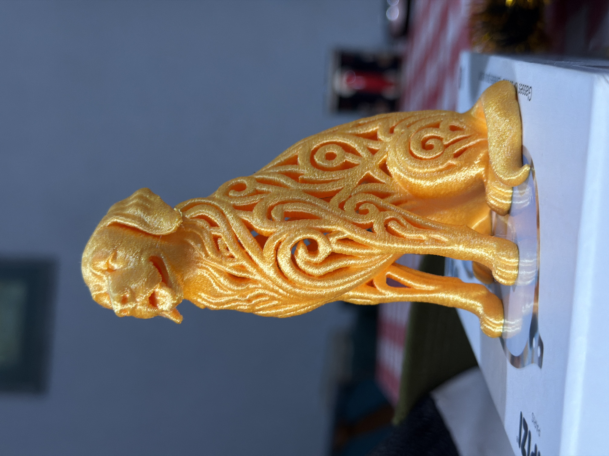 A 3D-printed, stylized, golden-colored dog figurine is the central object. The dog is depicted in a standing position, with intricate swirl patterns forming its body. It is sitting on a white box. A blue-gray wall serves as the backdrop. Other blurred objects are present, including what appears to be a picture frame, holiday-themed decor, and possibly a red and white patterned item. Location is not discernible from the image.