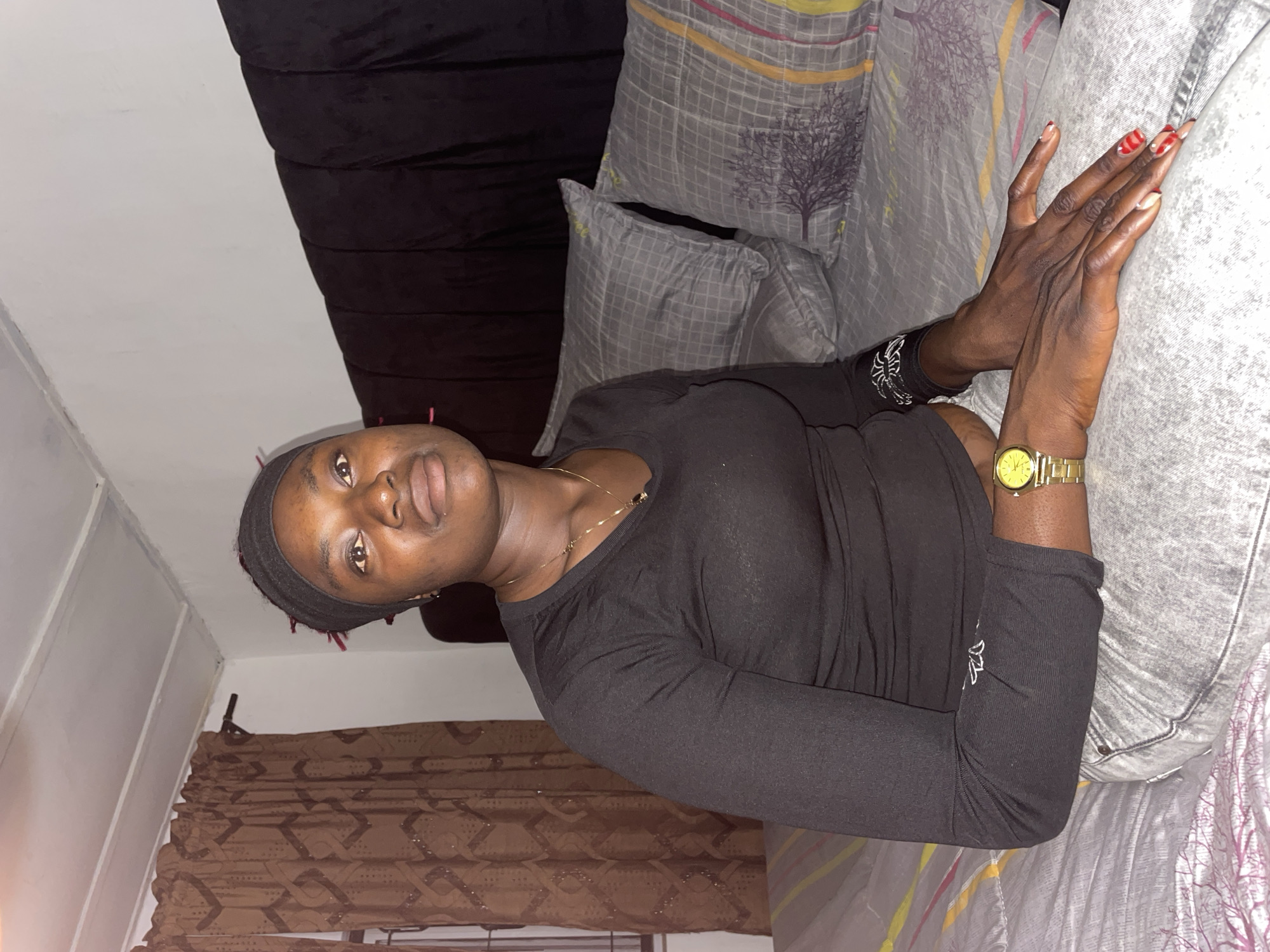 The scene is located in a bedroom within the city of Jos, Nigeria. A dark-skinned individual, appearing to be a young adult female, is seated upright on a bed. She is wearing a black long-sleeved shirt and light grey jeans. A black headband is worn on her head, with a few red strands visible underneath. A thin gold-colored necklace with a small dark pendant hangs around her neck. A gold-toned wristwatch is present on her left wrist. Her fingernails exhibit a red and white painted design, including red patterns on the tips. Her gaze is directed slightly upwards and to the right of the frame, with a neutral facial expression. Her hands are resting on her lap or jeans.
The bed features a grey bedspread and two pillows with a matching grid pattern, accented by yellow, pink, and purple stripes, and a dark tree silhouette design. Behind the pillows, a dark, padded headboard is visible. The room's walls are white, and the ceiling is white with visible structural seams or panels, showing some discoloration. To the left, a window fitted with metal security bars is partially obscured by brown curtains. The curtains display a repetitive dark geometric pattern and subtle shimmering details. The ambient lighting suggests an indoor setting, with illumination primarily from the front of the subject.