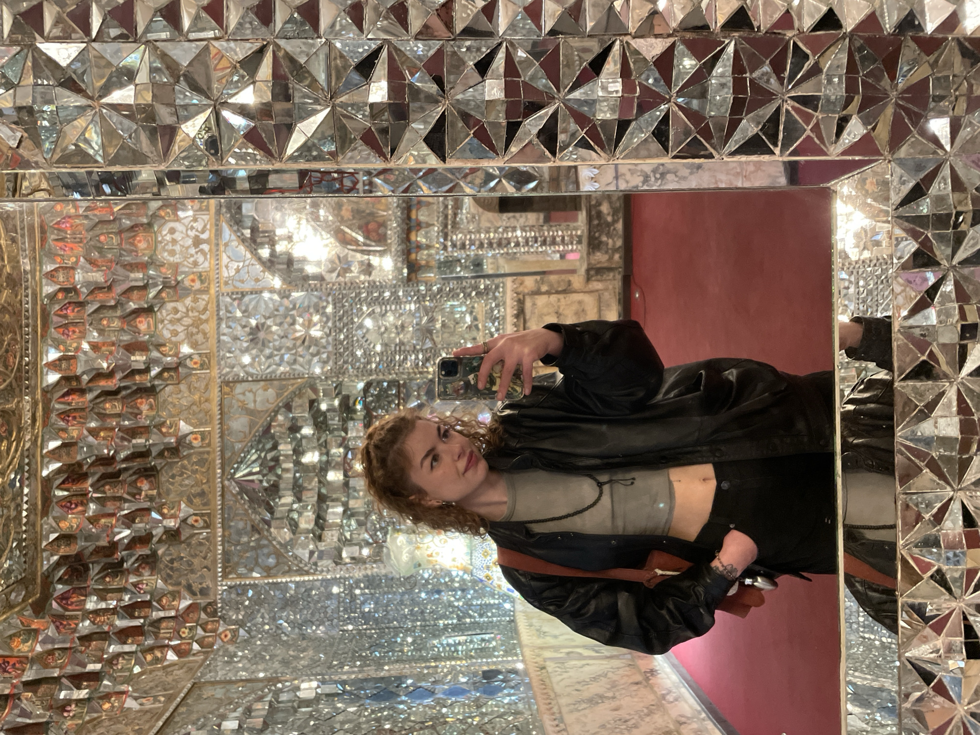 The image, rotated 90 degrees counter-clockwise, depicts a young woman taking a mirror selfie in a lavishly decorated room. The setting is characterized by an intricate mosaic of mirrors, known as "Aine-kari" or "Shishe-kari" (mirror work), a style common in traditional Persian and Georgian architecture, consistent with the location context of Tbilisi, Georgia.
The subject, a woman with reddish-brown, curly hair, is shown from the chest up. She is wearing a light gray cropped top, revealing her midriff, and a black leather jacket. A dark, possibly beaded, necklace is visible around her neck, and she wears a ring on her right hand. A brown strap, likely from a bag, crosses her chest. She holds a smartphone, possibly an iPhone, with a patterned case, to capture her reflection. Her expression is a calm, soft smile as she looks towards the camera.
The mirror reflecting the woman is framed by an ornate border composed of numerous triangular and star-shaped mirror pieces, creating a geometric and glittering pattern. The background, visible through and beyond this mirror, reveals more of the elaborate room. It is a dazzling display of various mirrored surfaces, some cut into small, faceted shapes, and others possibly adorned with gilded or painted details. A prominent deep red color, likely from a wall or floor covering, is visible in the reflected depths of the room, providing a warm contrast to the predominant silvery tones of the mirrors. The overall impression is one of intricate artistry and a shimmering, kaleidoscopic environment. No legible text is visible in the image.