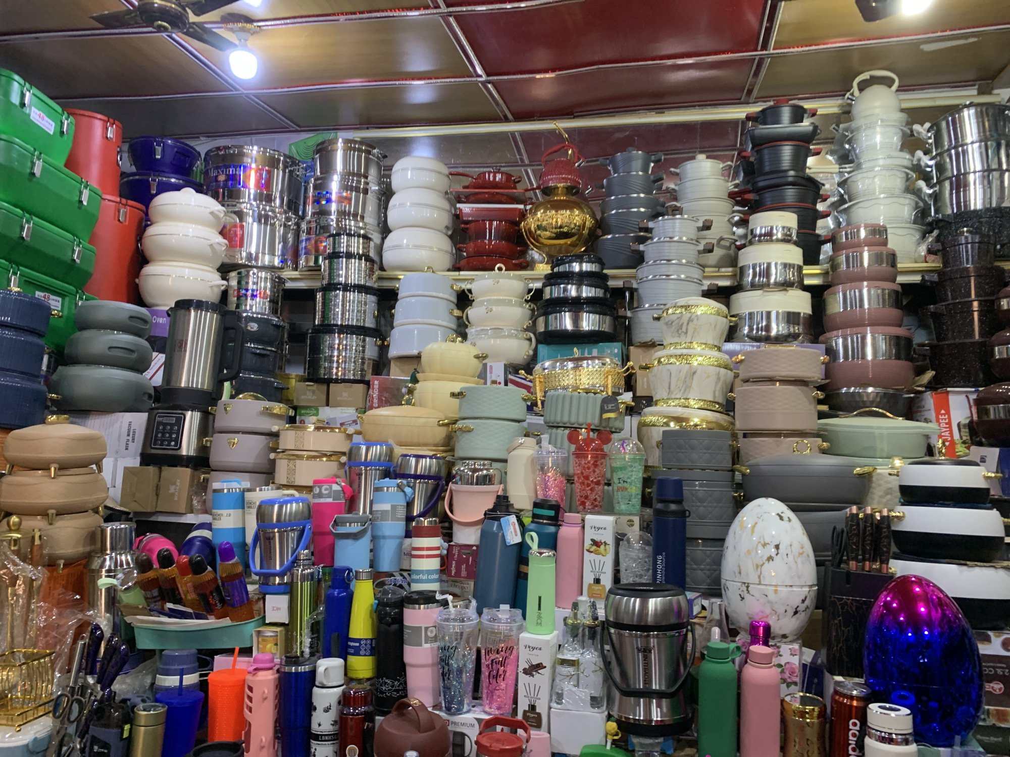 A cluttered store shelf is packed with stacked cookware sets, insulated food containers, thermoses, and assorted kitchenware. The merchandise is arranged densely, with a variety of colors, shapes, and materials. A ceiling with fluorescent lights and a fan are visible at the top. The foreground features items such as insulated water bottles, cooking utensils, and other smaller kitchen accessories. The OCR reveals that the location is the city of Jos, Nigeria.