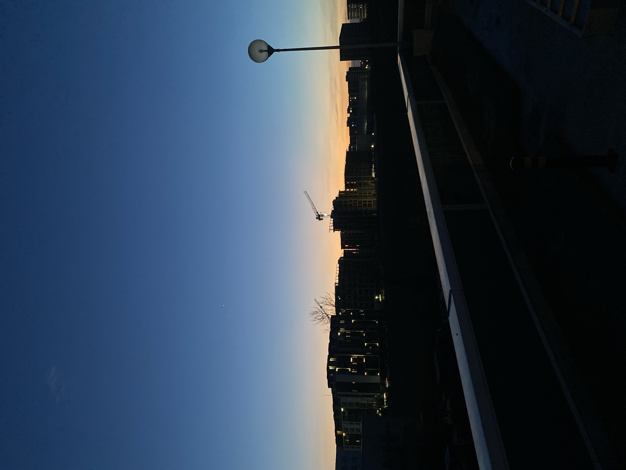 The image displays an urban landscape captured at twilight, presented as if rotated 90 degrees clockwise to show the horizon horizontally.
The sky, occupying the upper portion of the corrected image, exhibits a gradient from a deep, dark blue overhead to a lighter blue, transitioning into warm orange and yellow tones along the horizon. A single, bright star or planet is faintly visible in the upper left quadrant of the sky.
Silhouetted against this colorful evening sky is the skyline of a city. Numerous buildings, many appearing to be in various stages of construction with exposed frameworks and scaffolding, extend across the midground. A prominent construction crane stands tall among these structures, indicating ongoing urban development. Some finished buildings display illuminated windows, casting small glows against the darkening facades. A small, bare tree is also visible amongst the buildings.
In the left foreground of the correctly oriented image, a modern street lamp with a spherical light fixture stands on a tall pole, extending into the twilight sky. To the right, a dark railing or barrier runs diagonally, suggesting the photograph was taken from an elevated vantage point such as a bridge, balcony, or rooftop overlooking the city.
No specific human activity is depicted; the scene is static and quiet, capturing the peaceful transition from day to night in an urban or suburban setting. The overall mood is serene and contemplative. There is no visible text within the image. The setting is consistent with a developing urban area, such as Burgas, Bulgaria.