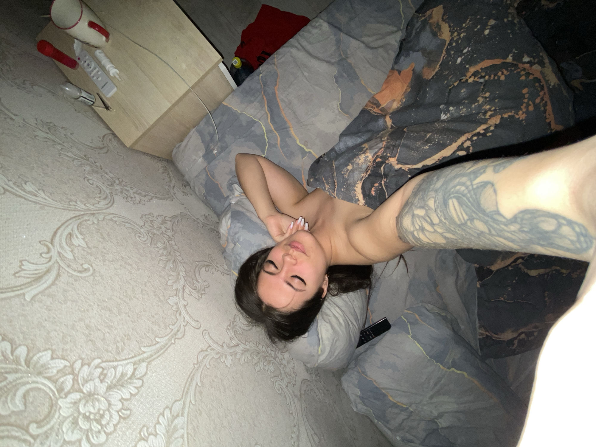 A selfie captures a young woman lying on her back on a bed in what appears to be a bedroom in Chaikovsky, Russia. Her eyes are closed, and she has dark hair and long, light-colored nails. She is topless, with her arms resting above her head, showcasing a large, intricate dark tattoo sleeve on her left arm. A black remote control rests on the light grey patterned duvet near her head.
The bed features two distinct duvet patterns: one is light grey with subtle wavy lines, and the other, partially pulled back, is a darker grey/black with striking gold and orange marbled designs. The room's wall to the left is covered with ornate beige or light grey wallpaper featuring elegant floral and damask patterns.
To the upper left, a wooden nightstand holds several items: a white mug with a red handle, a red cylindrical object, a white remote control with the brand 'LEDETTY' visible, another white cylindrical item, and a small metal clip. A white cable is also visible on the nightstand. Beyond the nightstand, on what appears to be the floor, a dark red fabric item and a dark bottle with a yellow cap are present.
The photograph is taken from a slightly elevated angle, with an extended arm, also tattooed with an intricate design, visible in the foreground, indicating a selfie. The low light suggests it is nighttime or late evening. The woman's closed eyes and relaxed pose convey a sense of calm or sleep.