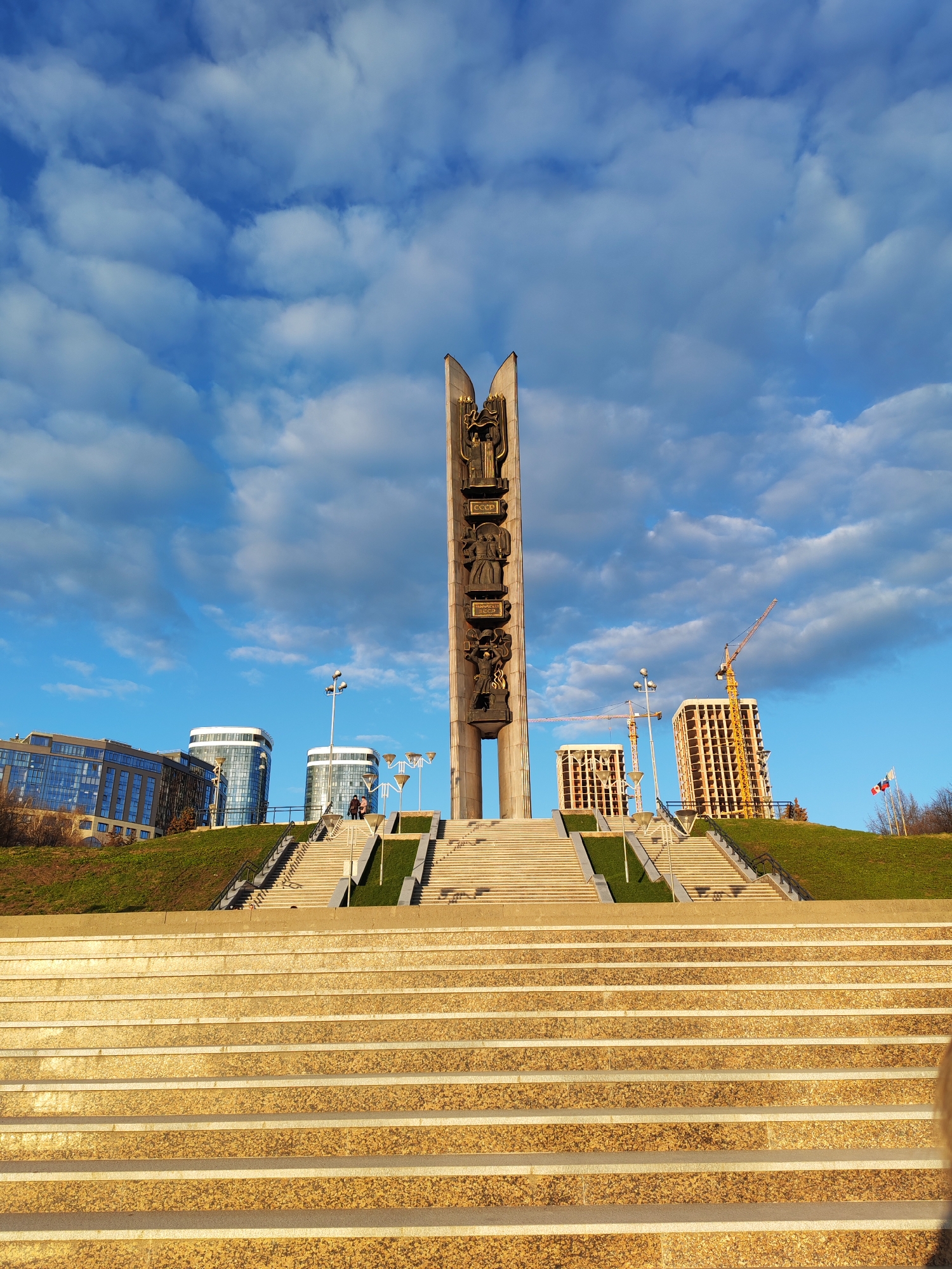 The image shows a monument in Staryi Senteg, Russia, viewed from a low angle at the base of a large set of stairs leading up to it. The monument is tall and rectangular, with bronze bas-reliefs depicting figures and inscriptions, the word "CCCP" visible. It has a central gap and stands on a green hill. Several sets of stairs, separated by green strips, lead to the top where people are walking. Behind the monument and the hill, there are modern buildings, some under construction with cranes. The sky is bright blue with fluffy white clouds. The foreground is dominated by the wide, shallow steps leading up to the monument. Flags on the right of the monument are waving in the wind. The image is taken in daylight.