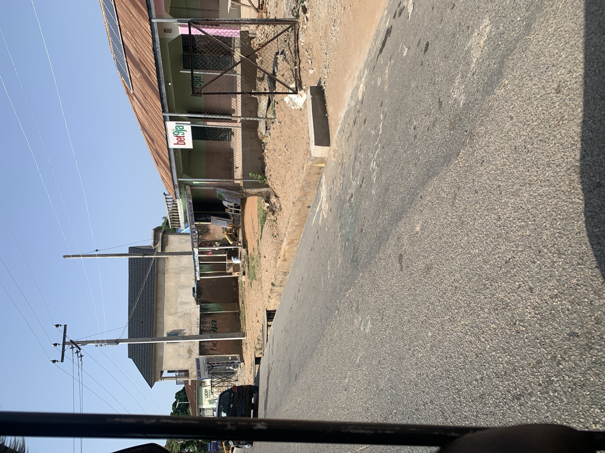 In a wide-angle shot taken from a vehicle, a street scene unfolds under a clear blue sky. A dark grey asphalt road occupies the foreground and midground, with a subtle curve to the right. A concrete curb runs along the edge of the road, separating it from a sandy embankment. On the embankment, a dark-colored sedan is parked.
Behind the embankment, several buildings are visible. To the left, a two-story building with a dark pitched roof stands. Adjacent to it, a single-story structure painted in light green and pink features a sign with the "bet9ja" logo. This building appears to be a shop, with a fence in front of it. Several other shopfronts with various signs are also visible, including "LUCKY BOOKSHOP". A utility pole with wires stands in front of the buildings, and solar panels are installed on the roof of the "bet9ja" shop. The environment appears somewhat undeveloped, with exposed sand and scattered debris. The time of day is likely morning or afternoon, given the bright sunlight and clear skies.