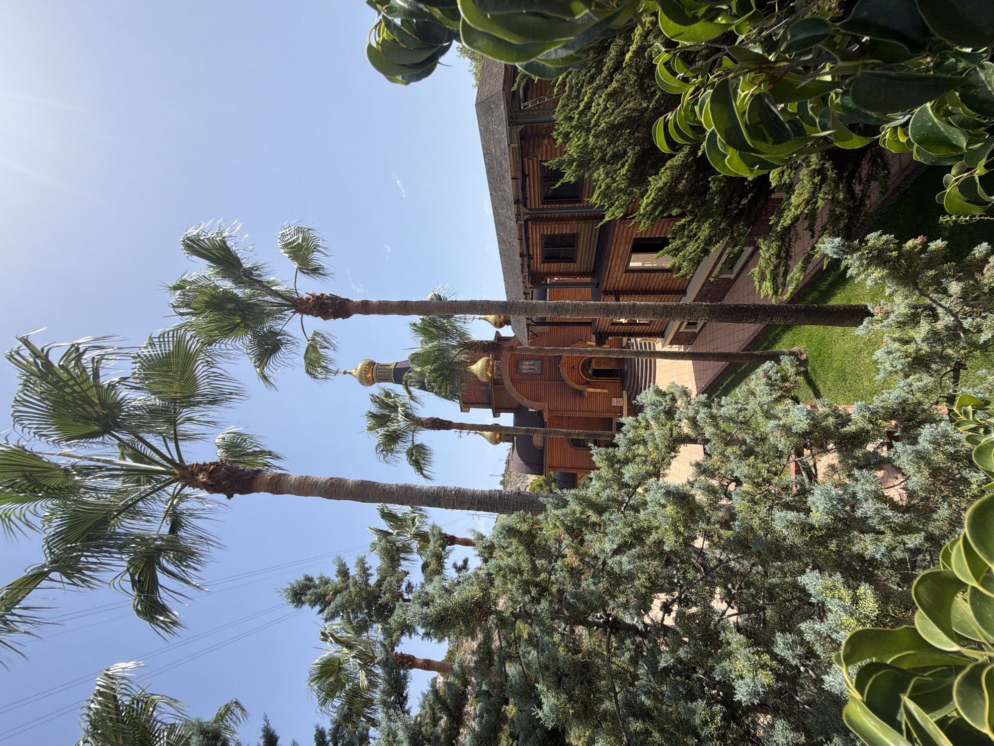 This image depicts the Russian Orthodox Church of San Miguel Arcángel in Altea, Spain. The church, constructed primarily of wood, features traditional onion domes topped with gold crosses. It is surrounded by lush greenery, including several palm trees and evergreen trees. The scene is set against a clear blue sky on a sunny day. The architecture suggests a Mediterranean or tropical climate, consistent with Altea's location on the Costa Blanca. The image captures the serene and picturesque atmosphere of the church grounds.