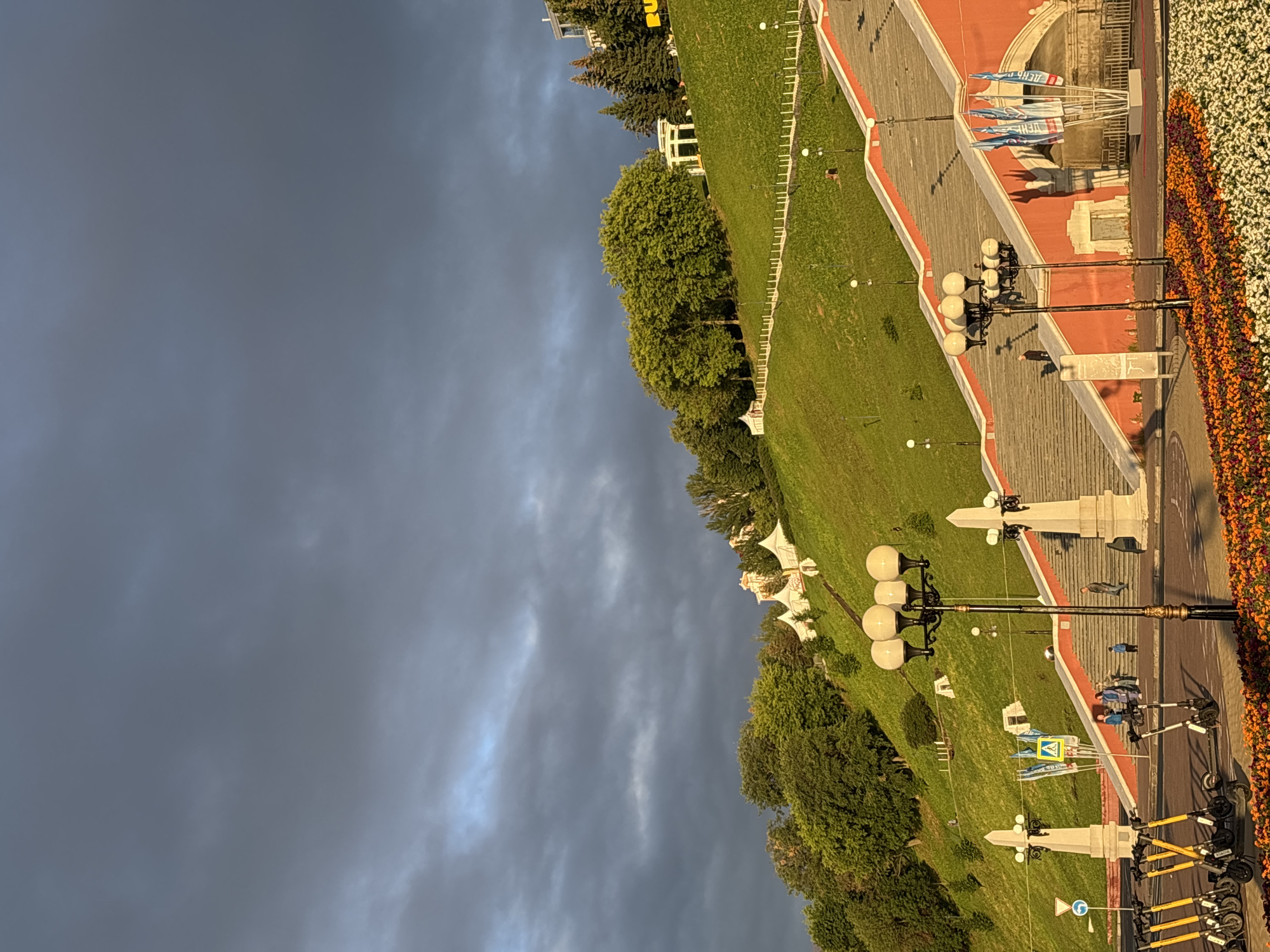 This is a low-angle, eye-level, medium shot of the Chkalov Stairs in Nizhny Novgorod, Russia, on a cloudy day with shafts of sunlight breaking through. The foreground shows a sidewalk with several electric scooters parked and a flowerbed with orange and purple flowers. The Chkalov Stairs, a prominent landmark in the city, are visible on the right, with their red walls and wide concrete steps ascending the green hillside. Several people are walking up and down the stairs. Flags are displayed near the bottom of the stairs.
The middle ground features a lush green hillside dotted with trees and several white tents, possibly for an event. A white gazebo-like structure is visible near the top of the hill, surrounded by trees. The sky is filled with dark, dramatic clouds, with patches of lighter sky and sunlight illuminating parts of the hillside and the clouds themselves. The overall impression is one of an outdoor public space with historical architecture and natural elements, under a dynamic sky.