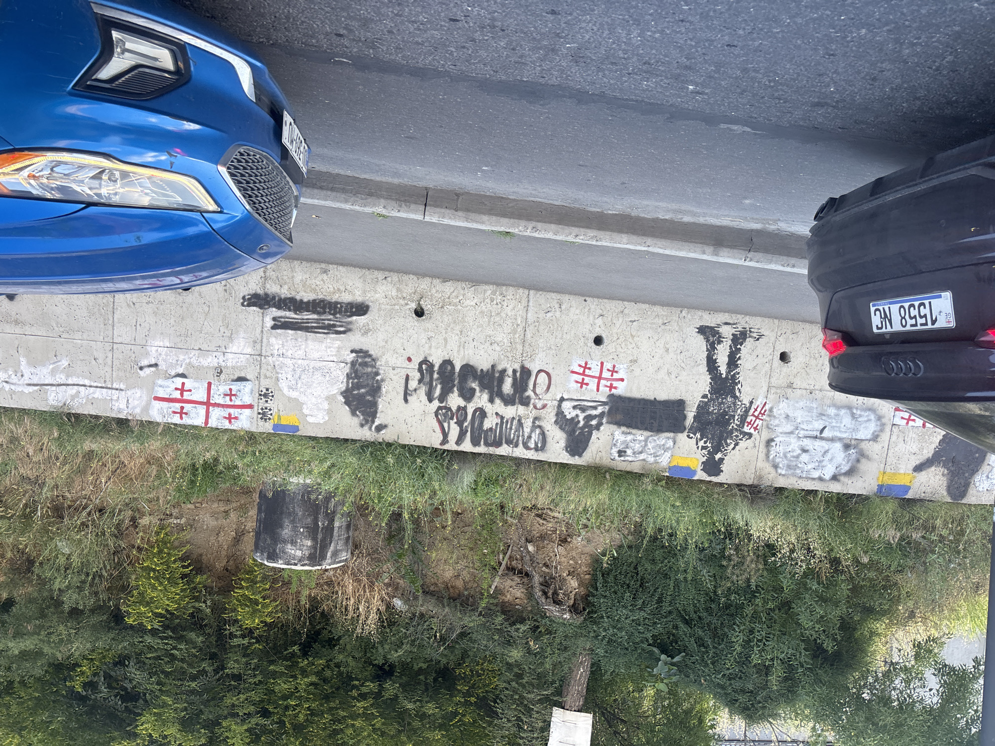In an outdoor urban environment, likely Tbilisi, Georgia, based on the license plate on the black Audi, a concrete wall along a slope is covered in graffiti and political symbols. The wall, which appears to be a retaining wall, is adorned with spray-painted images of figures, some in silhouette, and what appear to be national flags. Specifically, there are two Ukrainian flags (blue and yellow) and at least two Georgian flags (white with red crosses). One of the graffiti figures is a soldier-like silhouette holding a flag. Text in what appears to be Georgian script is also present in black spray paint. The area behind the wall is densely overgrown with trees and vegetation, with exposed soil and roots on the slope. A large, cylindrical concrete structure, possibly a pipe or storage container, is visible on the right side of the image, partially obscured by foliage. In the foreground, two cars are parked on a paved road. The rear of a black Audi with a Georgian license plate reading "1558 NC" is visible on the left, and the front of a bright blue car is visible on the right. The road has a concrete curb separating it from the wall. The scene is illuminated by daylight, suggesting it is daytime.