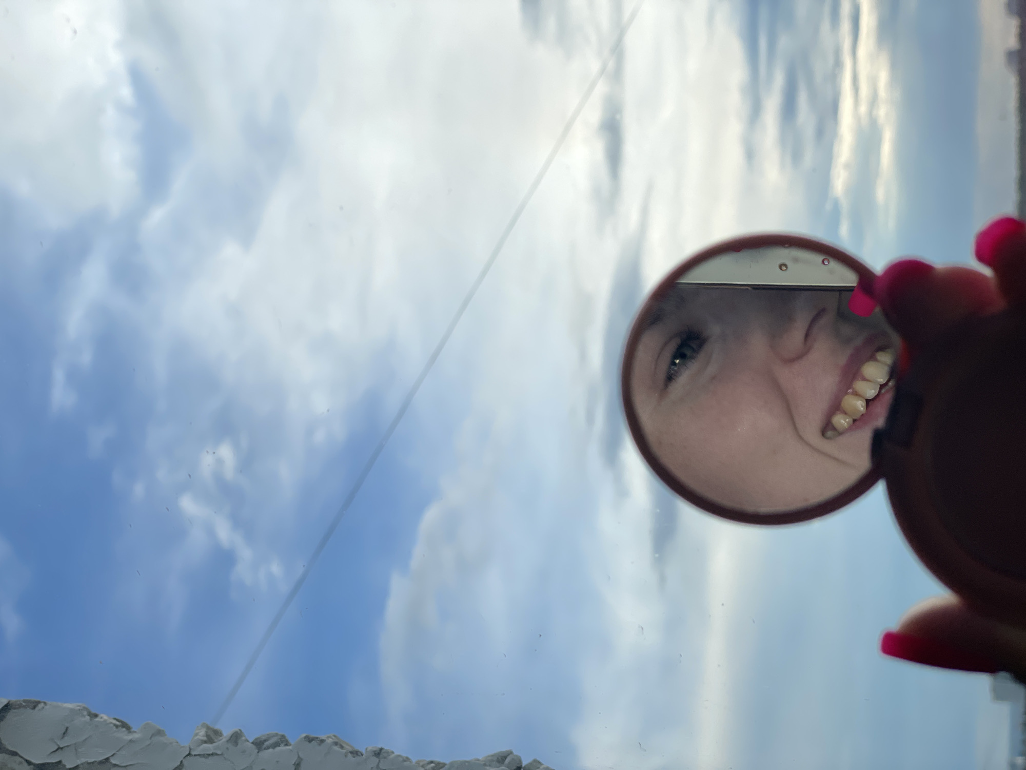 The image, oriented vertically, captures an upward perspective towards a bright daytime sky. The central subject is a person's cheerful face, reflected in a small, round compact mirror. The mirror is held by a hand, identifiable by fingers with vibrant pink nail polish. The reflection shows a distinct blue-green eye and a wide, toothy smile, conveying a joyful or amused expression.
The background features a vast expanse of clear blue sky adorned with scattered white cumulus clouds, indicating bright and pleasant weather. A faint, thin diagonal line crosses the sky, possibly a scratch on a window or a distant wire. In the lower left section, a textured, light-colored surface, resembling a building's facade or a window ledge, is visible. The image captures a mirrored self-portrait against the sky, set in Izhevsk, Russia. No visible text is present.