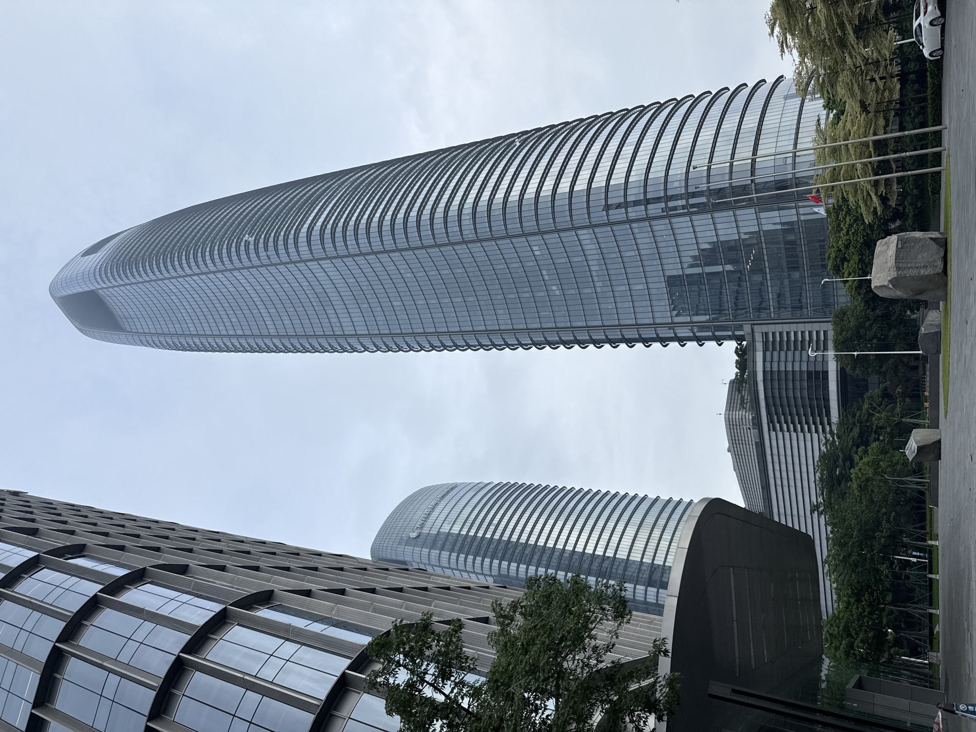 The image depicts a low-angle view of modern skyscrapers in Haizhu District, China. On the left, a tall building with a textured grey facade and large rectangular windows rises against a pale, overcast sky. To its right, a second, more rounded skyscraper with a reflective glass facade features horizontal banding and curves inwards towards its top, which has a distinctive curved opening. In the background, a lower, multi-level building with a stepped, ribbed facade is visible, surrounded by lush green trees. The foreground shows a paved plaza with a large, rough-hewn rock. Three tall, slender flagpoles stand in front of the rounded skyscraper, one of which has a red and white flag. A white car is parked on the right side of the image. The overall scene conveys a sense of urban modernity and architectural scale.