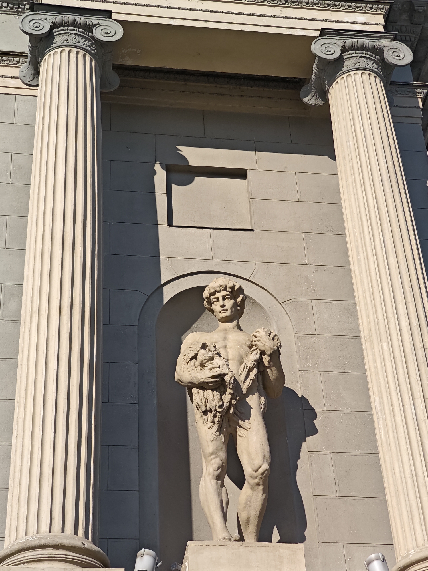 A stone sculpture of a muscular male figure is centrally located within an alcove of a building. The sculpture depicts the figure holding a bundle of what appears to be foliage or flowers. The architectural facade features large, fluted columns on either side of the alcove. The building's exterior is constructed of stone blocks. The lighting suggests a sunny day, casting prominent shadows from the columns and the sculpture onto the wall. The scene appears to be an exterior shot of a public building in Moscow, Russia, likely a museum or a government structure given the classical architectural style and the presence of decorative sculpture.