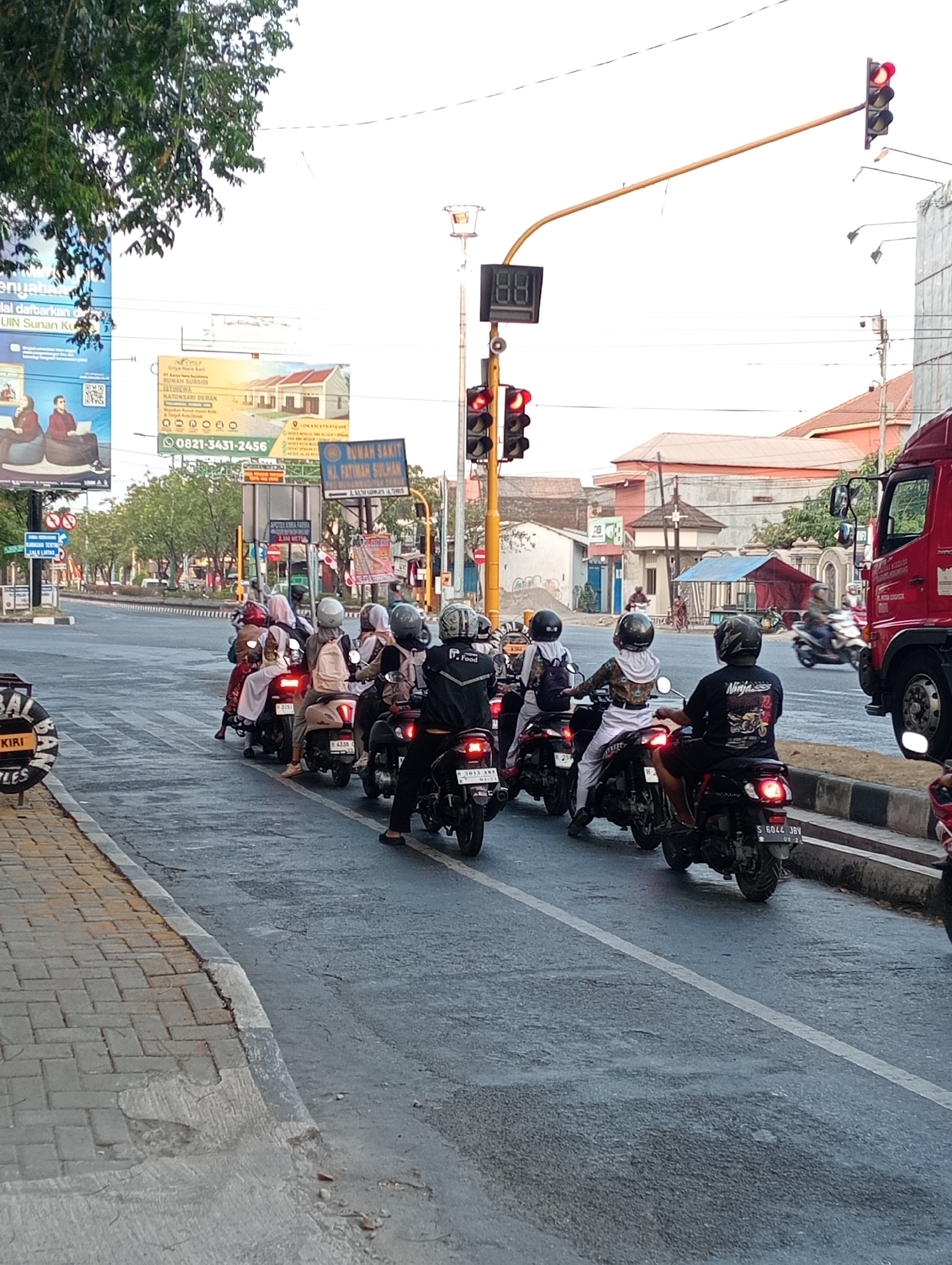 A group of motorcyclists are waiting at a traffic light in Demak, Indonesia, as indicated by the "DEMAK" text on a billboard in the background. The scene depicts a busy street with multiple lanes of traffic. The motorcyclists, some wearing helmets and face coverings, are lined up on their bikes. A red truck is visible in the background, along with various buildings and billboards. The traffic light is red, and a digital timer shows "88," suggesting a countdown to the light change. The weather appears to be overcast.