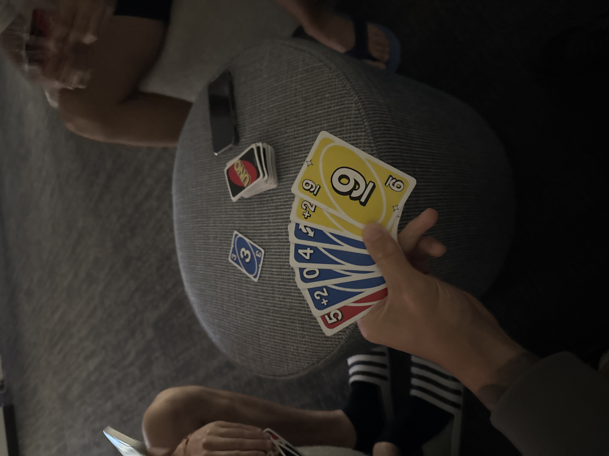 The image captures a casual, indoor setting, likely in the evening or a dimly lit room. The primary activity is playing Uno.
Two individuals are partially visible. One person's hands are prominent in the foreground, holding a fanned hand of Uno cards, revealing a yellow "9" (two copies), a blue "+2", a blue "0", a blue "4", and a red "5". This person is wearing black and white striped socks and a dark top. Another person's arm, torso, and foot, clad in a blue flip-flop, are visible on the left, resting on a light-colored surface; their movement is slightly blurred.
The game is being played on a grey, textured, upholstered pouf or stool. On this surface, a stack of face-down Uno cards bearing the "UNO" logo is visible, alongside a single face-up blue "3" card, indicating the discard pile. A dark-colored smartphone lies face-down next to the card stack. The flooring appears to be carpeted. The lighting is artificial and low, contributing to a relaxed atmosphere. No specific location cues are visible within the image itself beyond the indoor setting, although the context provided places it in Myrtle Beach, USA. The scene suggests leisure and social interaction.