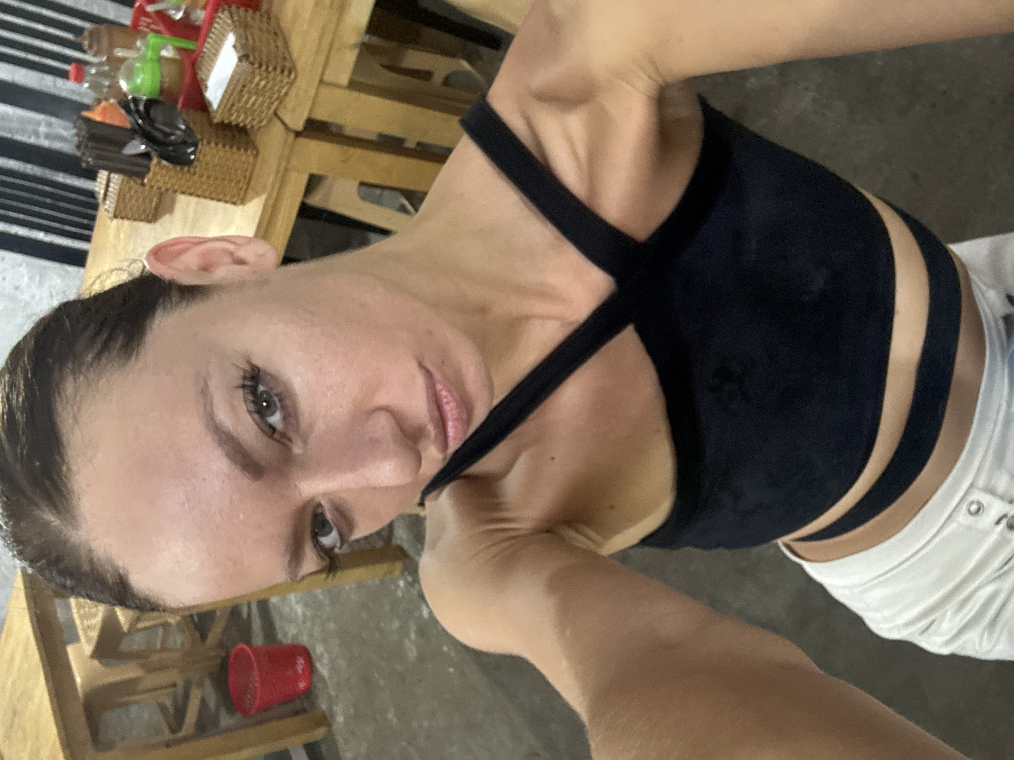 A woman is looking at the camera, wearing a black sports bra with cross straps and white shorts. The environment includes an indoor setting with wooden furniture, a red cup on a chair, and various items on a table including baskets and bottles. The location may be Đà Nẵng, Vietnam.