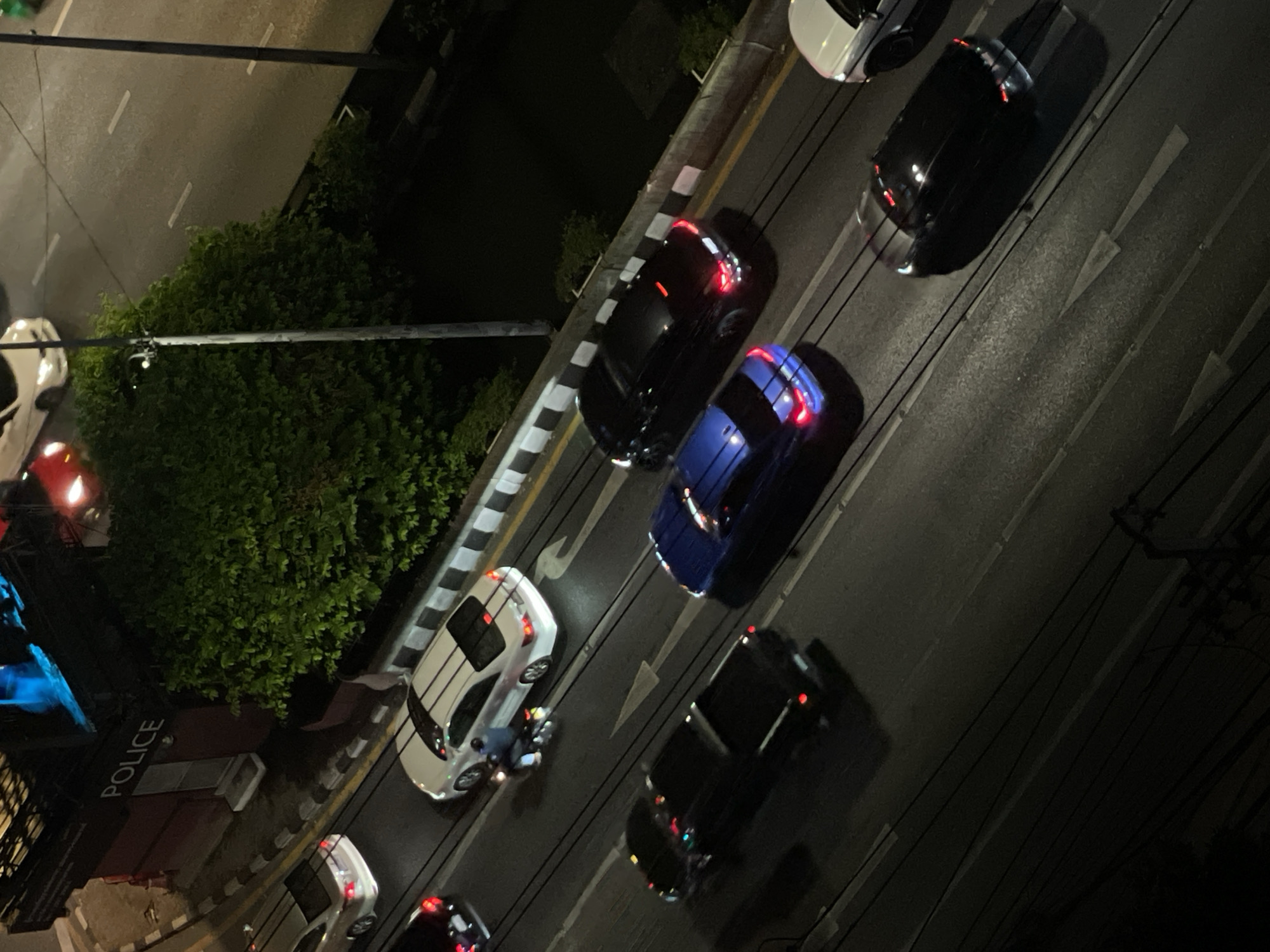 The image presents an overhead, angled view of a street scene at night in Bangkok, Thailand. The dark setting is illuminated by car lights and ambient street lighting, casting shadows from vehicles and overhead power lines.
The street appears busy with multiple vehicles. On the left side of the road, a white car is partially visible near the bottom, and another white car further up is accompanied by a motorcycle or scooter to its side. Adjacent to these, in the next lane, is a dark blue car, followed by several black and other dark-colored cars. Another white car is visible on the far right lane. Many of the cars have their tail lights on, glowing red.
The road surface is asphalt, marked with distinct white lane lines and large white arrow symbols indicating the direction of traffic flow. A curb with alternating black and white stripes, along with a yellow line, separates the road from a lush strip of green trees and bushes lining the left side of the street. Numerous dark power lines stretch across the upper portion of the image.
In the lower-left section, a building is partially visible, featuring a dark facade with the word "POLICE" inscribed in prominent white capital letters. The overall scene depicts active nighttime traffic in an urban setting.