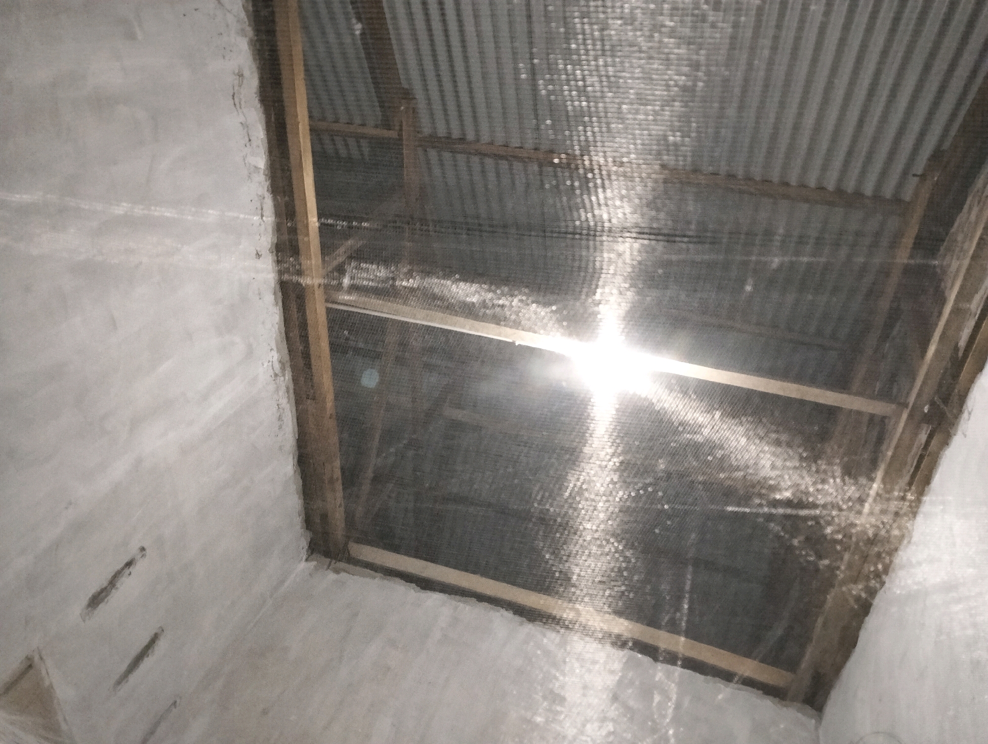 The image depicts the interior of a room with a corrugated metal roof and a white concrete wall. There is a bright light source in the center of the image, shining through a mesh material that covers a portion of the ceiling. The mesh is supported by wooden beams. The wall has several horizontal indentations and appears to be rough-textured. The scene is dimly lit, with the bright light creating a halo effect. The location is identified as Blang, Indonesia.