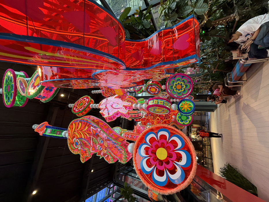 The image depicts a grand indoor public space within Iconsiam, Bangkok, Thailand. Overhead, a dense installation of large, vibrantly colored decorative elements hangs from a dark ceiling. These include numerous circular, flat ornaments featuring intricate floral patterns in red, pink, blue, yellow, and green. Interspersed are elongated, multi-tiered structures resembling stylized mythological figures or traditional lanterns, rendered in bright reds, oranges, and greens, with detailed patterns. A large, reflective, red, rectangular lantern-like structure dominates the right side of the frame.
The floor is a light-colored, reflective walkway. On the right, a row of three blue and red checkered benches is positioned alongside lush green foliage and plants, some adorned with string lights. Behind the greenery, a glass wall is partially visible. On the far left, a red escalator descends. In the background, a large illuminated screen with white text on a blue background is visible.
Four individuals are seated on the benches on the right, all engaged with their smartphones. In the mid-ground on the left, one male wearing a red long-sleeved shirt, dark pants, and a dark backpack walks away from the viewer along the walkway.