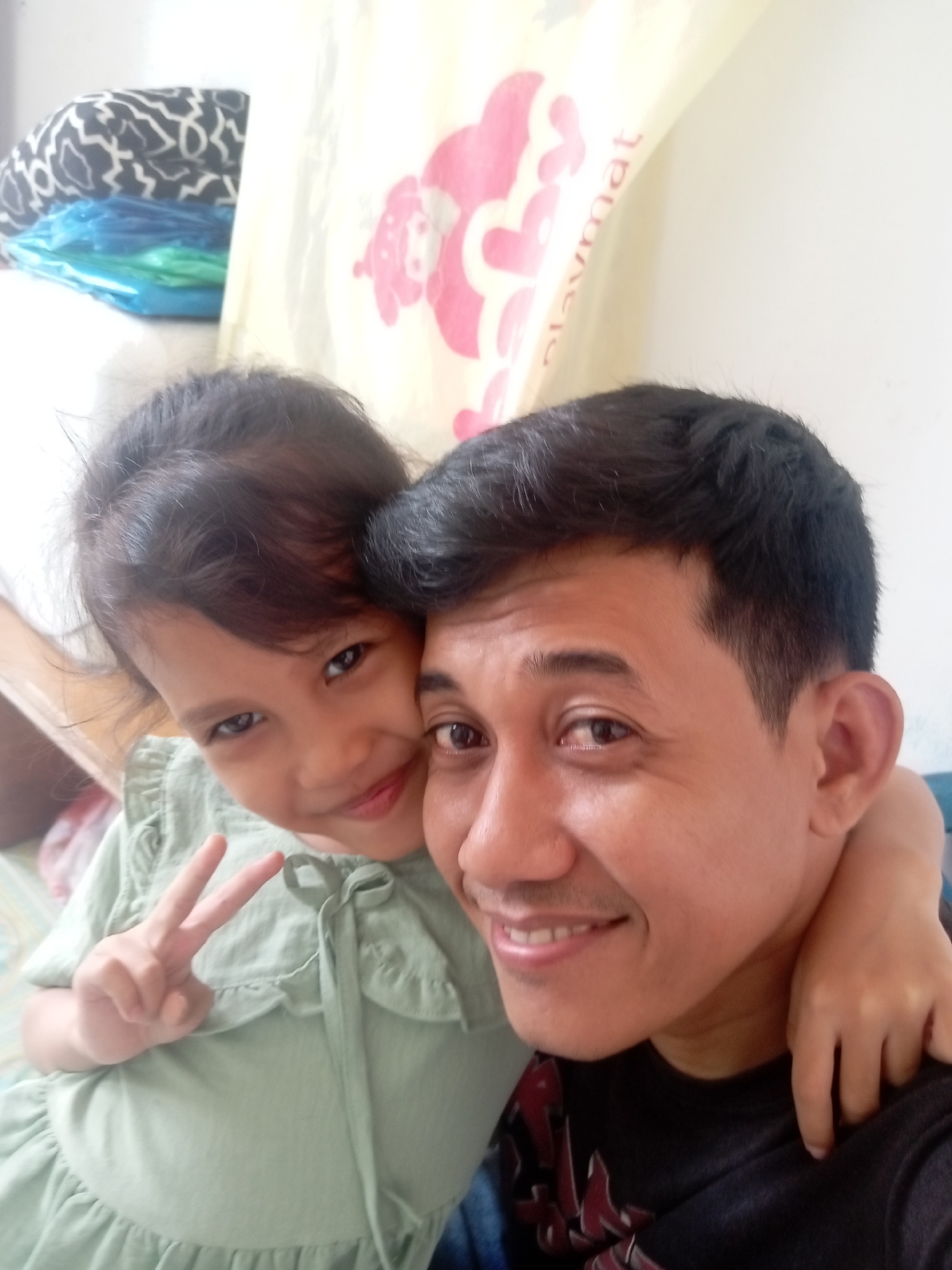 The image is a selfie taken indoors, likely in a residential setting in Batam, Indonesia. The foreground features a man and a young girl. The man, positioned on the right, has dark hair and is smiling, with his right arm around the girl's shoulders. The girl, on the left, is leaning into the man and making a peace sign with her left hand. She has dark hair and is wearing a light green dress. In the background, a white wall is visible, along with some blurred objects, including a patterned cushion and what appears to be a rolled-up playmat with pink graphics. The lighting suggests it is daytime.