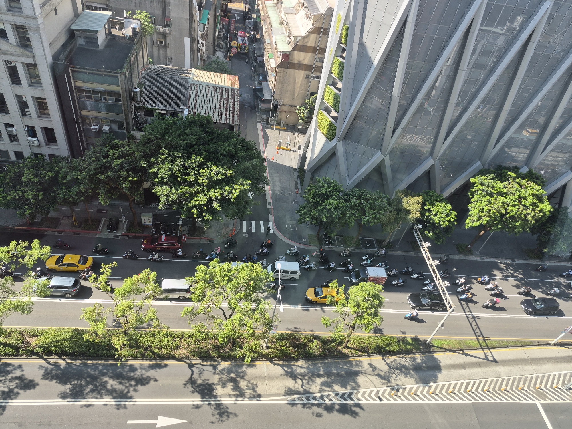 The image provides an aerial view of a street scene in Taipei, Taiwan, focusing on traffic and urban architecture. A multi-lane road runs horizontally across the lower portion of the frame, with a median strip of green shrubs and trees. A yellow taxi and several motorcycles are visible on the road, along with a few cars. The left side of the image features older, multi-story buildings with visible windows and air conditioning units, interspersed with lush green trees. On the right, a modern, angular building with a glass facade and integrated green planters dominates the background, reflecting the sky and surrounding environment. The street itself shows painted lane dividers and pedestrian crosswalks, indicating an active urban intersection. The overall impression is of a bustling city environment with a mix of old and new architectural styles and continuous vehicular movement.